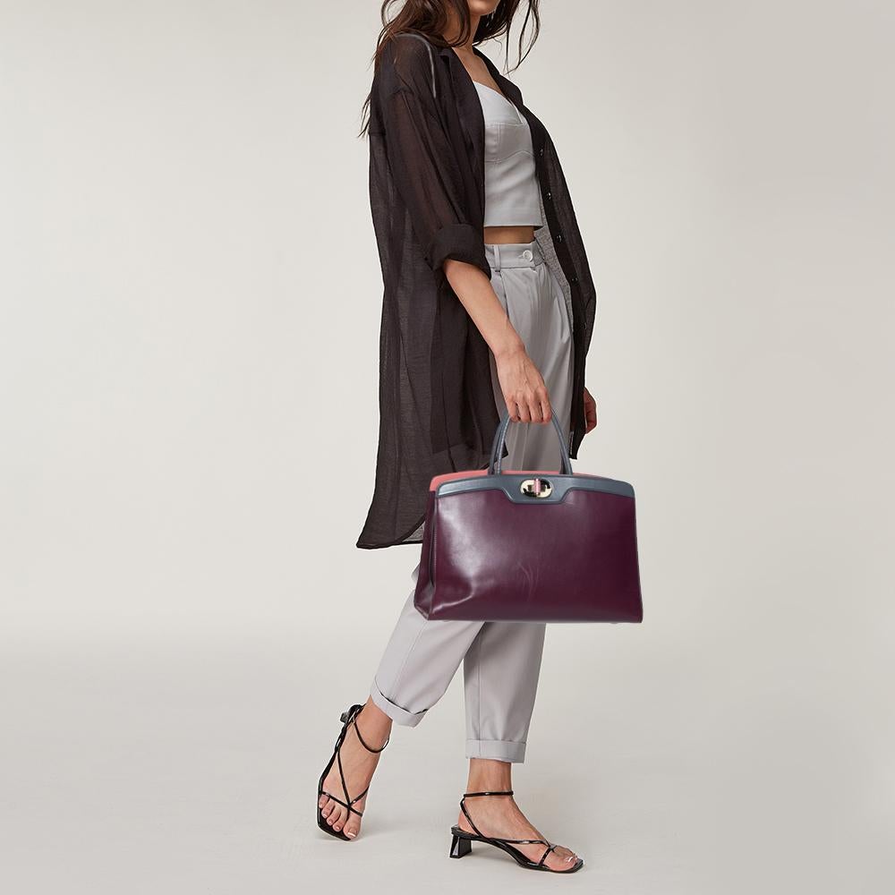 The sleek Icona tote from Bvlgari is made of burgundy & grey leather. A central zippered compartment separates the two open compartments. The lining has an open wall pocket and a tab through which the turn-lock secures the bag. The rolled handles