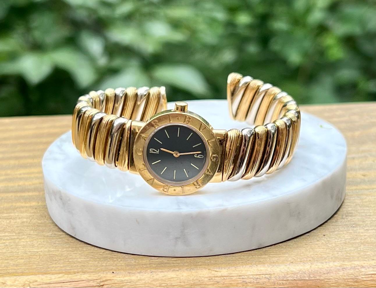 One 18 Karat yellow and white gold Bvlgari wristwatch bangle with a polished finish. The case is in yellow gold with a polished finish and has BVLGARI engraved around the bezel. Black dial with gold-tone hands. The band is two-tone gold and is 15mm