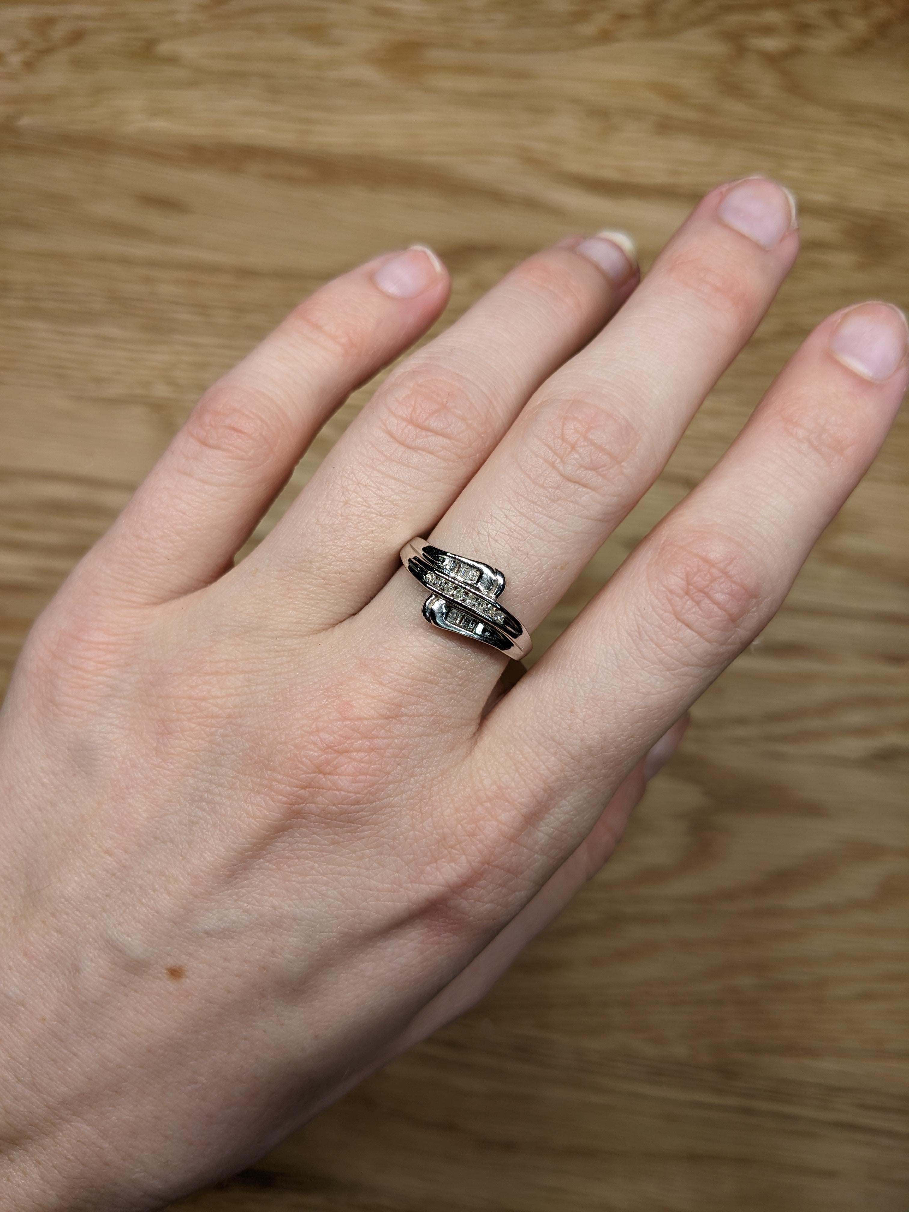 This isn't just a ring—it’s a conversation starter! The graceful bypass shank wraps your finger in polished 10K white gold, while round and baguette diamond accents add a burst of brilliance. Perfect for an engagement, anniversary, or a “treat
