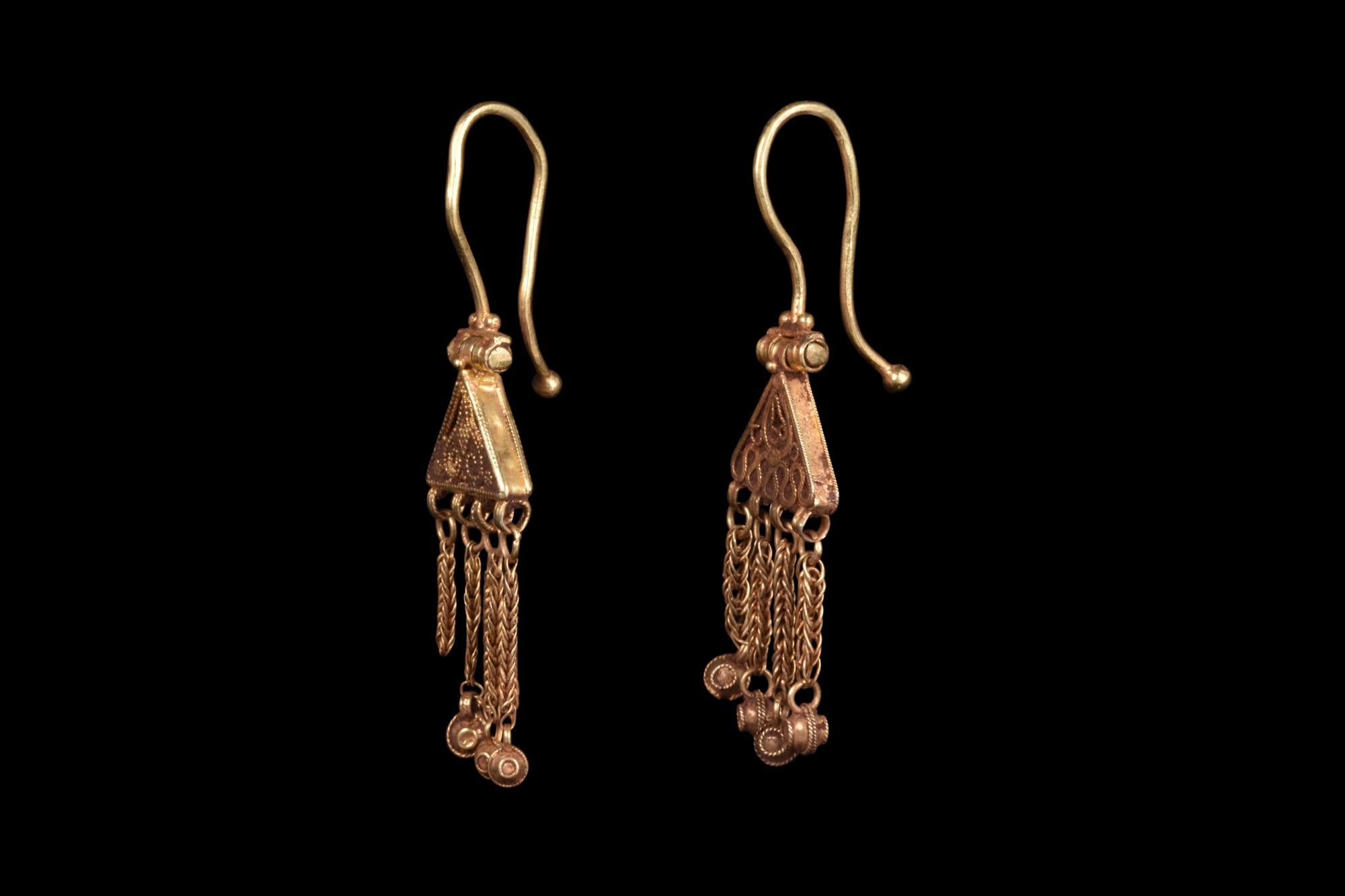 A stunning matched pair of Byzantine gold earrings, each comprised of a hinged and elongated hoop attached to a triangular-shaped plaque with an openwork filigree decoration. Suspended below are four chain dangles with intricately decorated gold