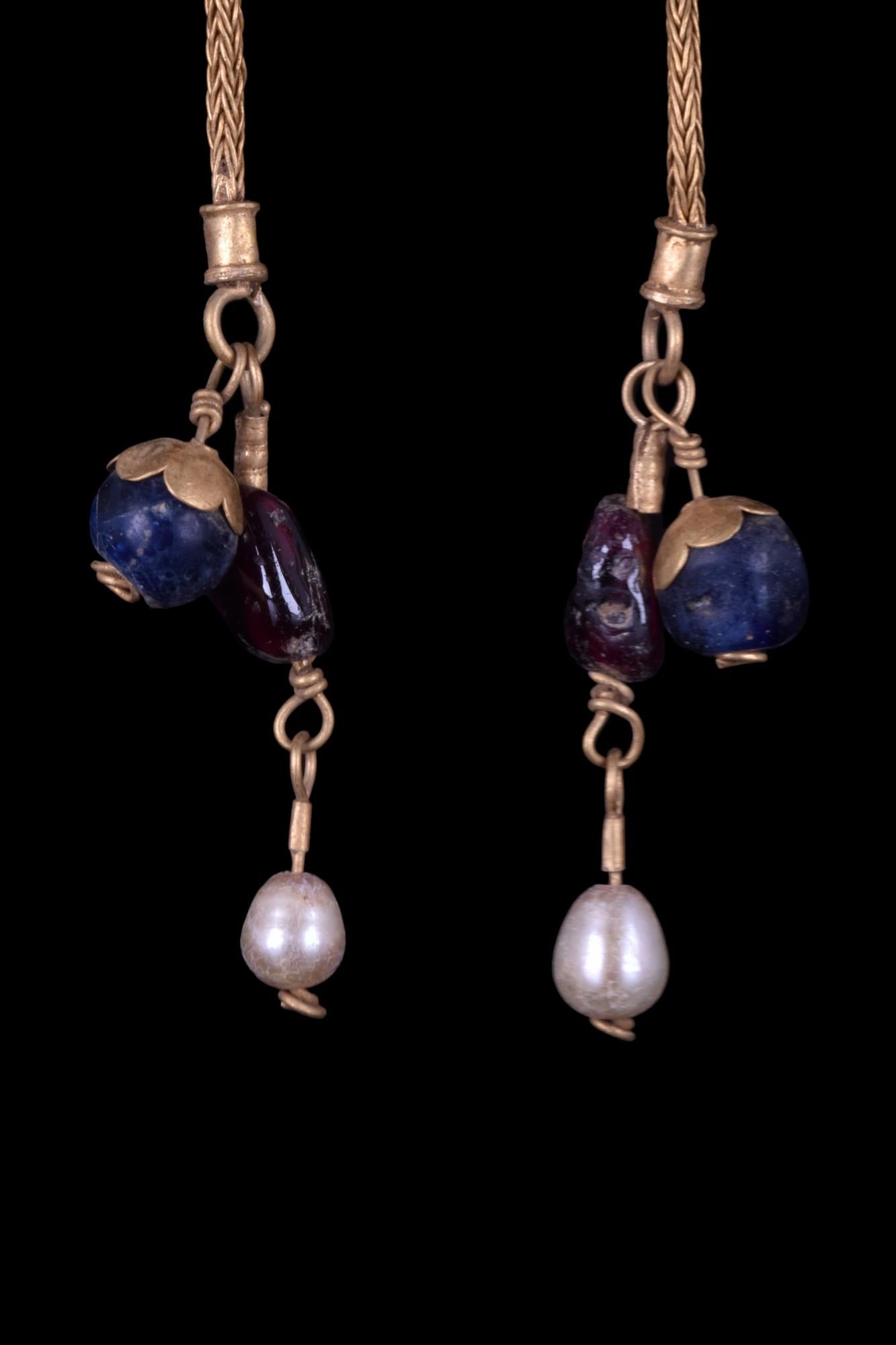 A matched pair of Byzantine gold earrings, each with a tapering hoop, hook-and-clasp closure, and a single elongated pendant. The pendant is composed of glass and pearl spherical beads, attached to a cord of woven gold wire. These beautiful pieces