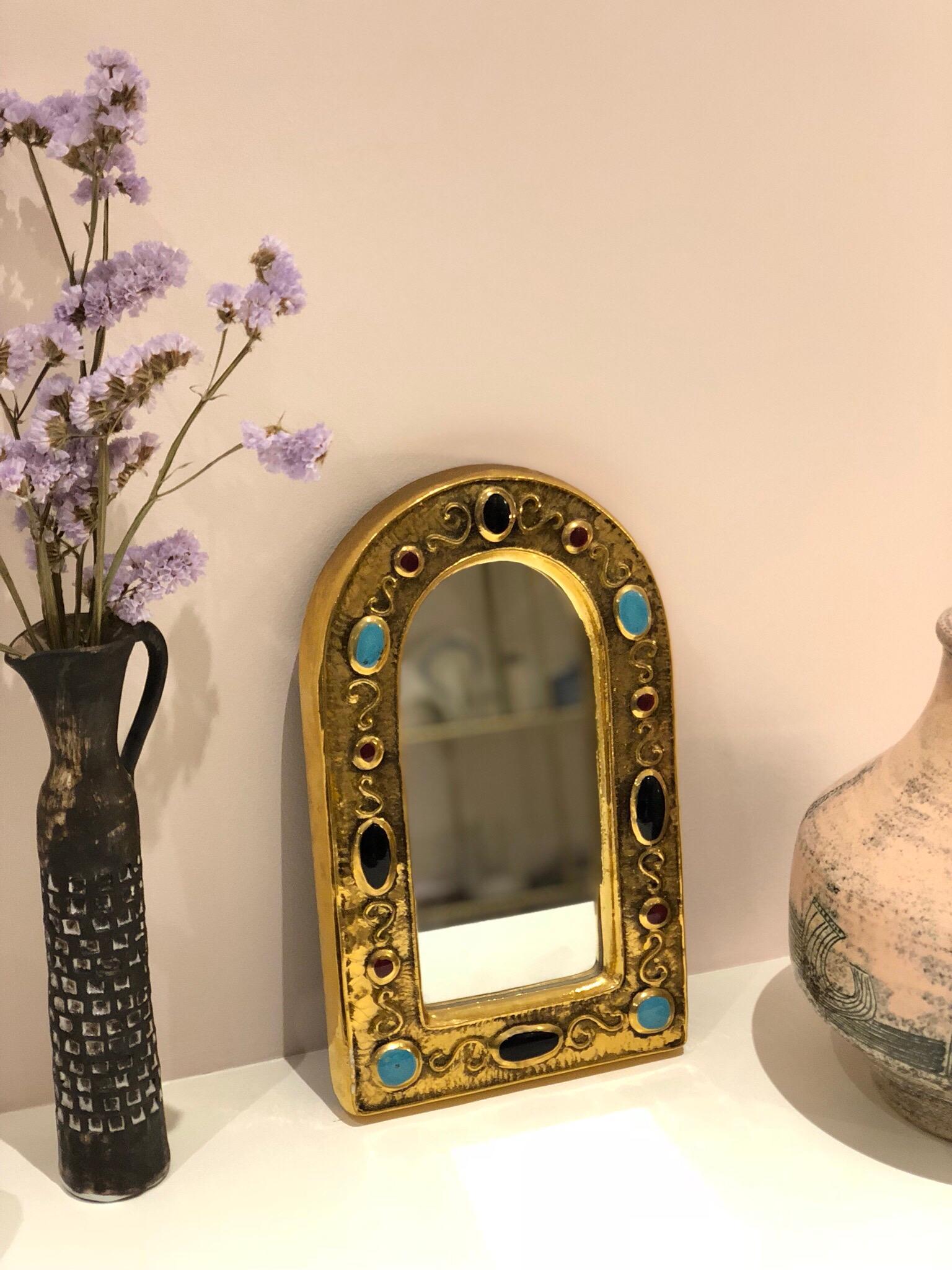 Inspired by Byzantine iconography, a gilded glazed ceramic wall mirror by François Lembo (circa 1960s - 70s). With enamelled ceramic cabochons in alternating colours of black, ruby red and turquoise, it is an unusually elegant piece of decorative