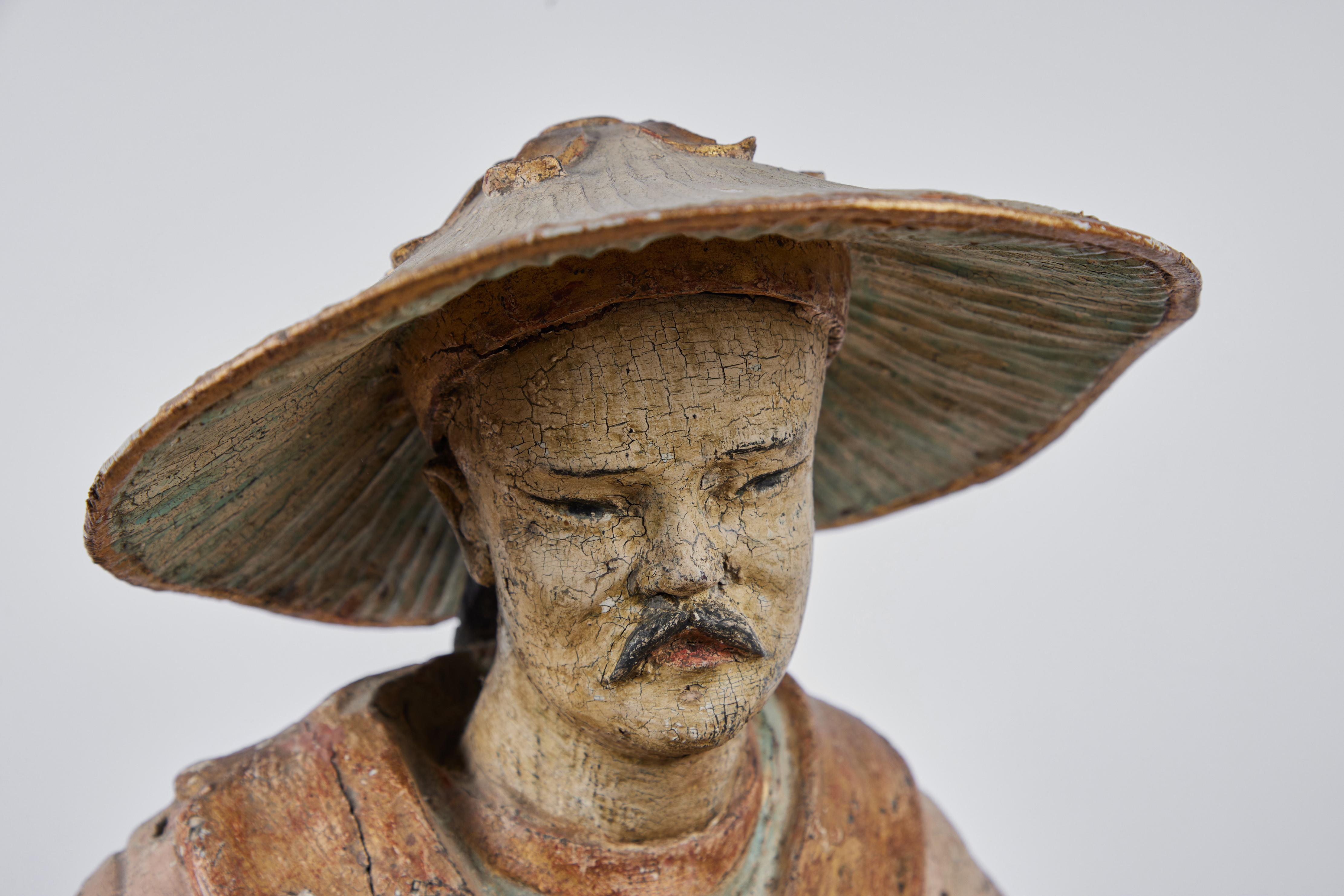 An elegant, turn-of-the-19th century, hand-carved, fully finished polychrome and parcel gilt statue of a Chinese wise man with a cascading braid beneath a a grand, bamboo hat capped by a foliate finial. He grasps both a scroll and a plumed fan. The