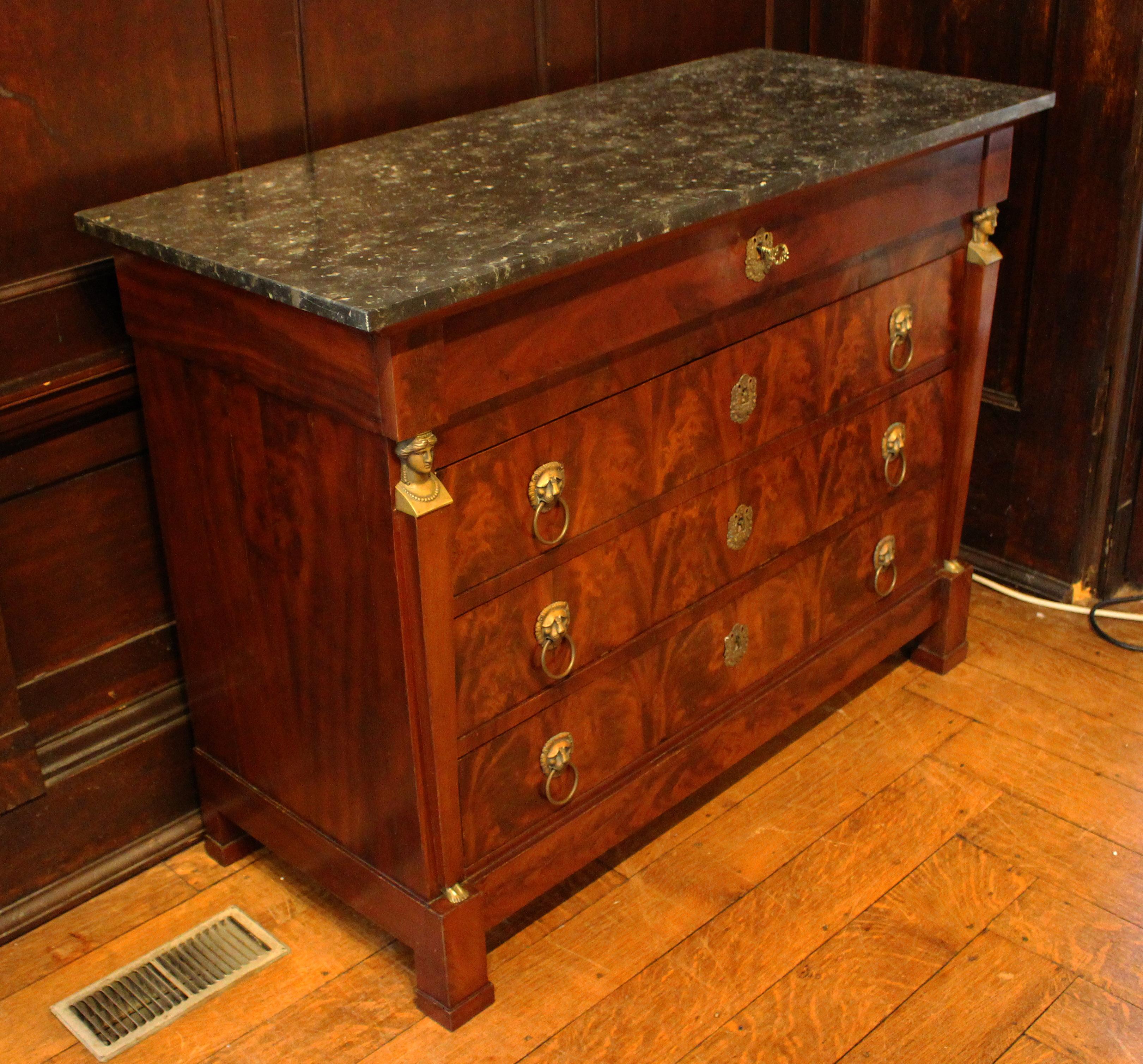 A French Empire marble top commode, circa 1820s-30s. Flame mahogany front flanked by pilasters featuring Grecian women with bronze busts & feet. Later ring turned lion mask pulls. Bronze escutcheons. Square straight feet ending in plinths. Oak