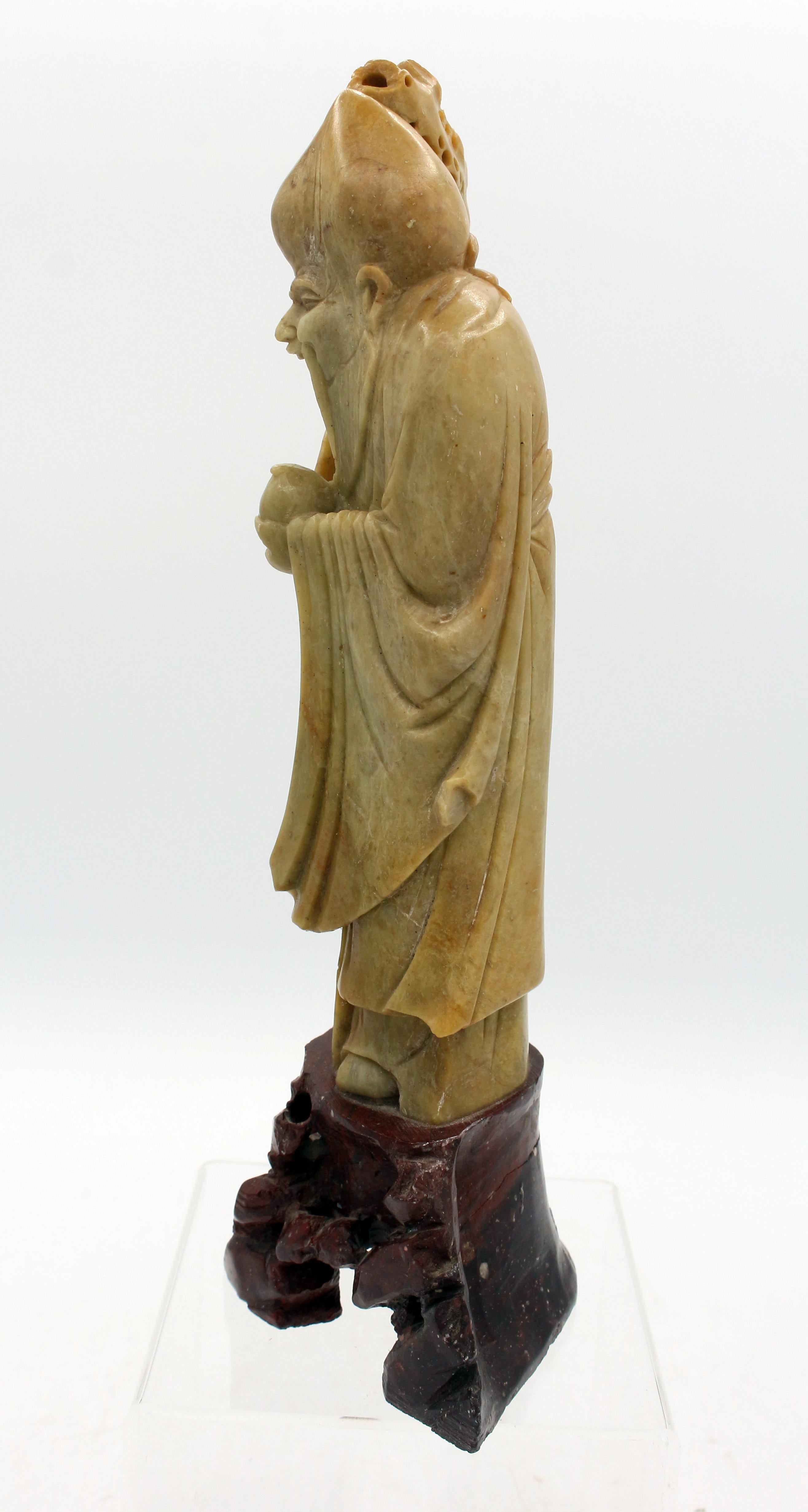 Figura in pietra ollare cinese del 1900 circa raffigurante Shouxing, dio della longevità. Ben scolpito con in mano una pesca e un lungo bastone. Su base affiorante scolpita in pietra ollare (vecchia rottura e riparazione visibile dall'interno).