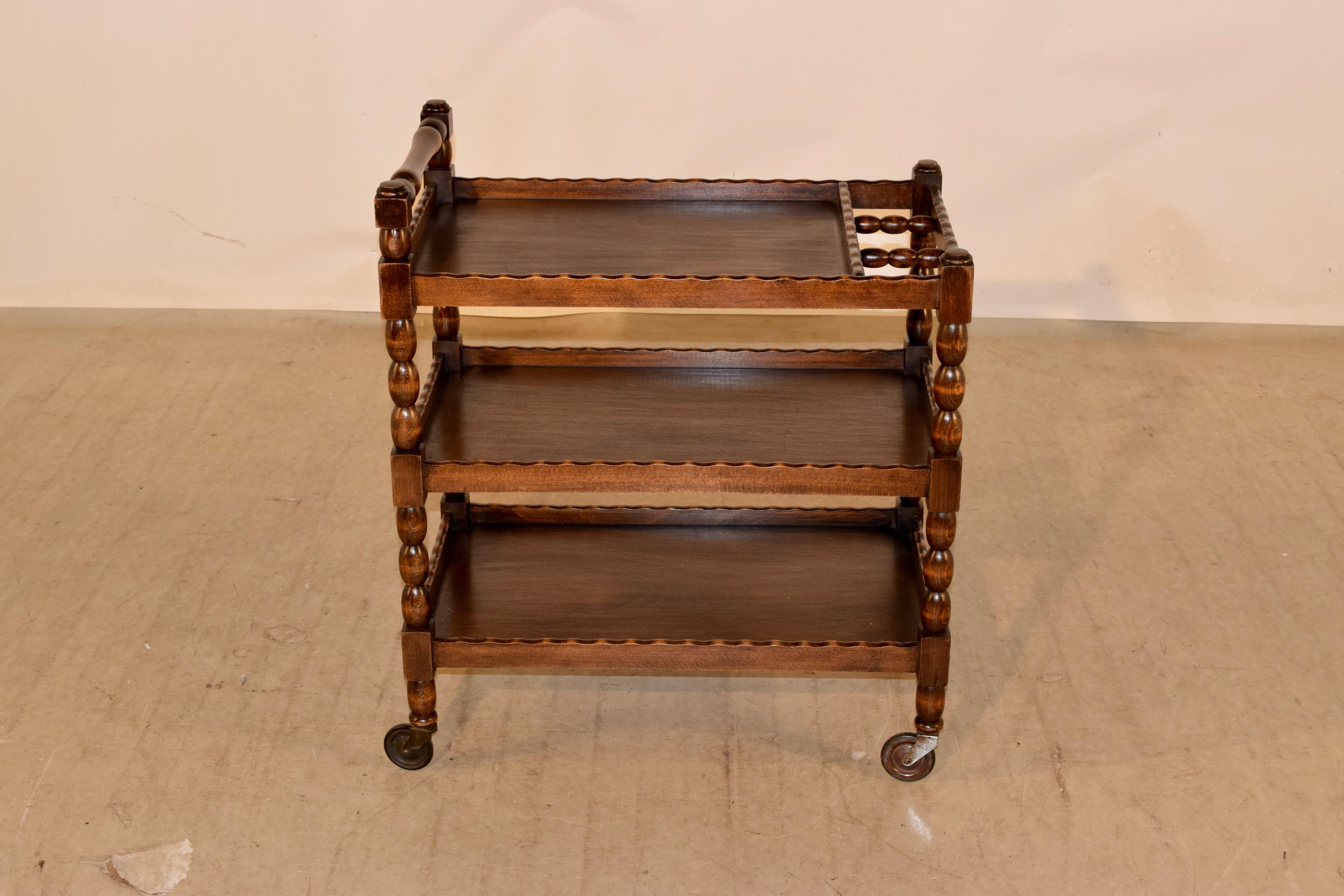 C. 1900 English oak drinks cart with a turned handle over three shelves which have scalloped galleries and hand turned shelf supports and feet. The cart is supported on what appear to be the original casters.