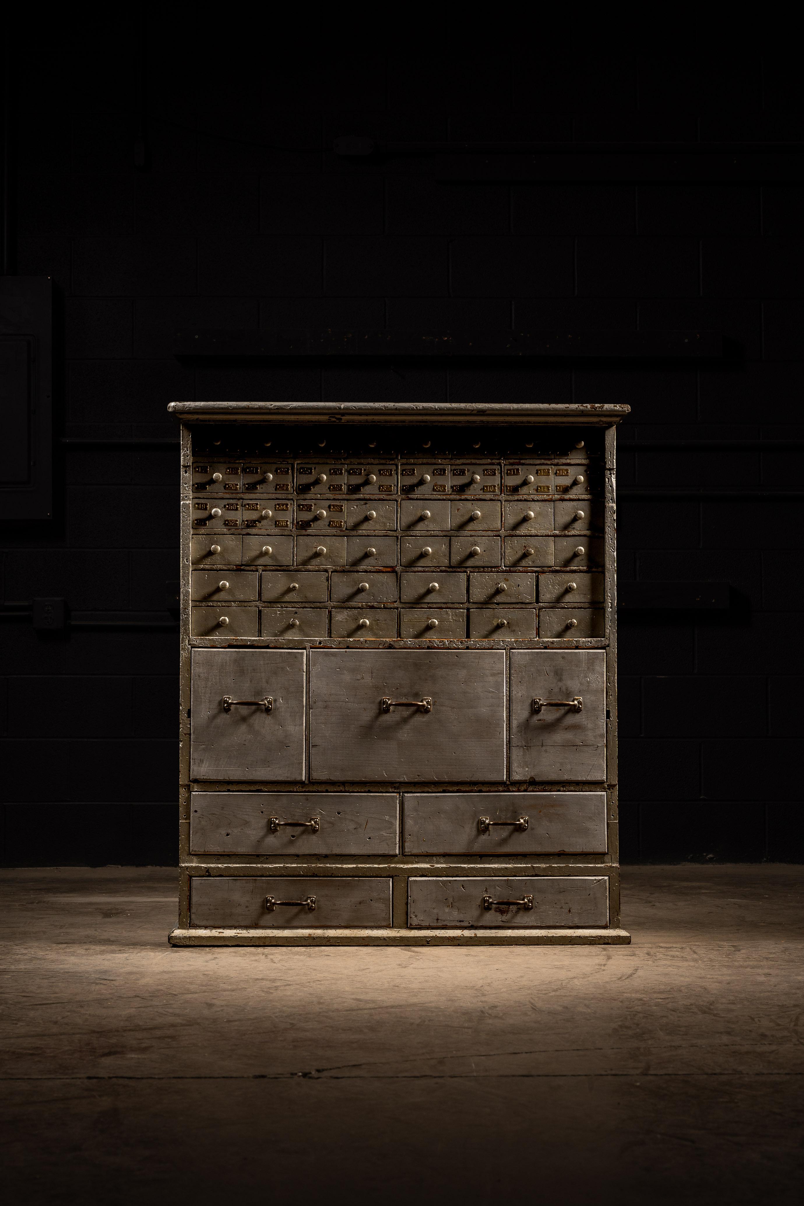 Industrial wooden cabinet with a silver and gray paint coat.
This piece features drawers of various sizes from small parts to general storage. Present on the smaller compartment drawers are brass number tags that count up to 60.

Structurally sound,