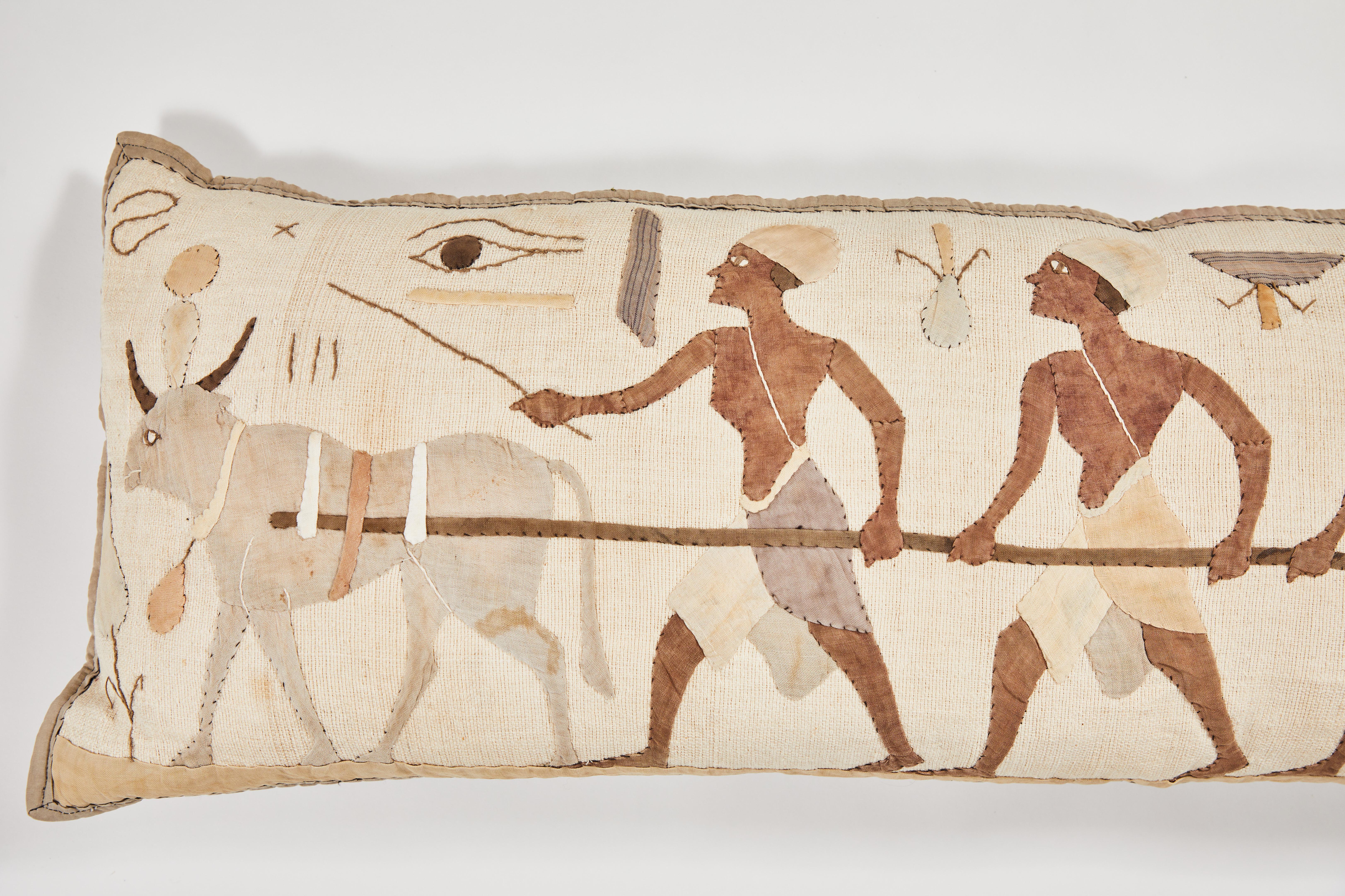 A 1920s textile with a hand-embroidered Egyptian scene in neutrals has been re-purposed and custom made into a long decorative bolster pillow for a sofa or bed. With running stitch symbols and appliquéd ox, toiling workers, falcon, eye of Horus,