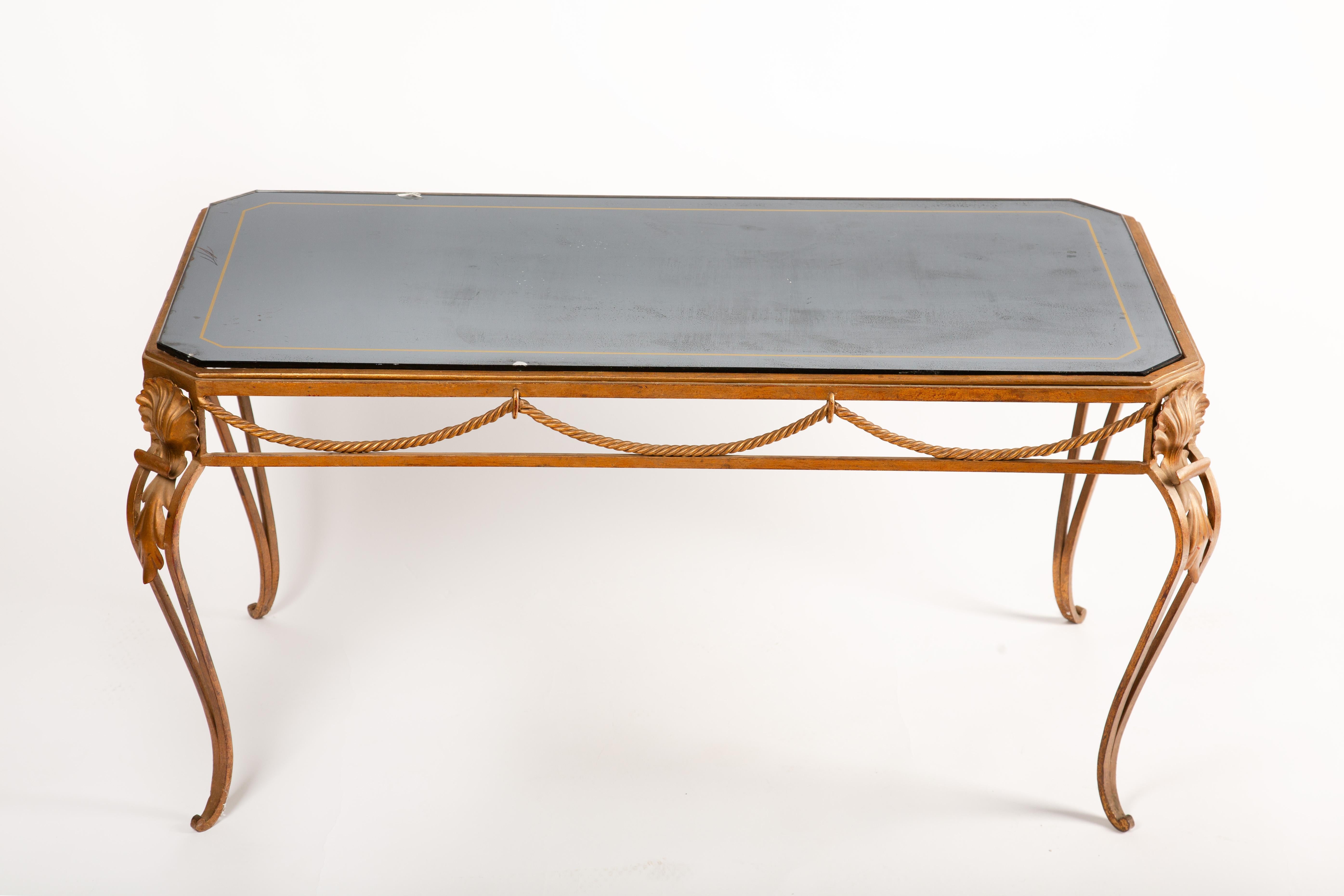 Cette élégante table basse française, datant d'environ 1940, présente une base en fer doré finement ouvragé avec des détails néoclassiques, notamment des guirlandes de corde, des motifs de coquillages et des pieds gracieusement enroulés. Le plateau