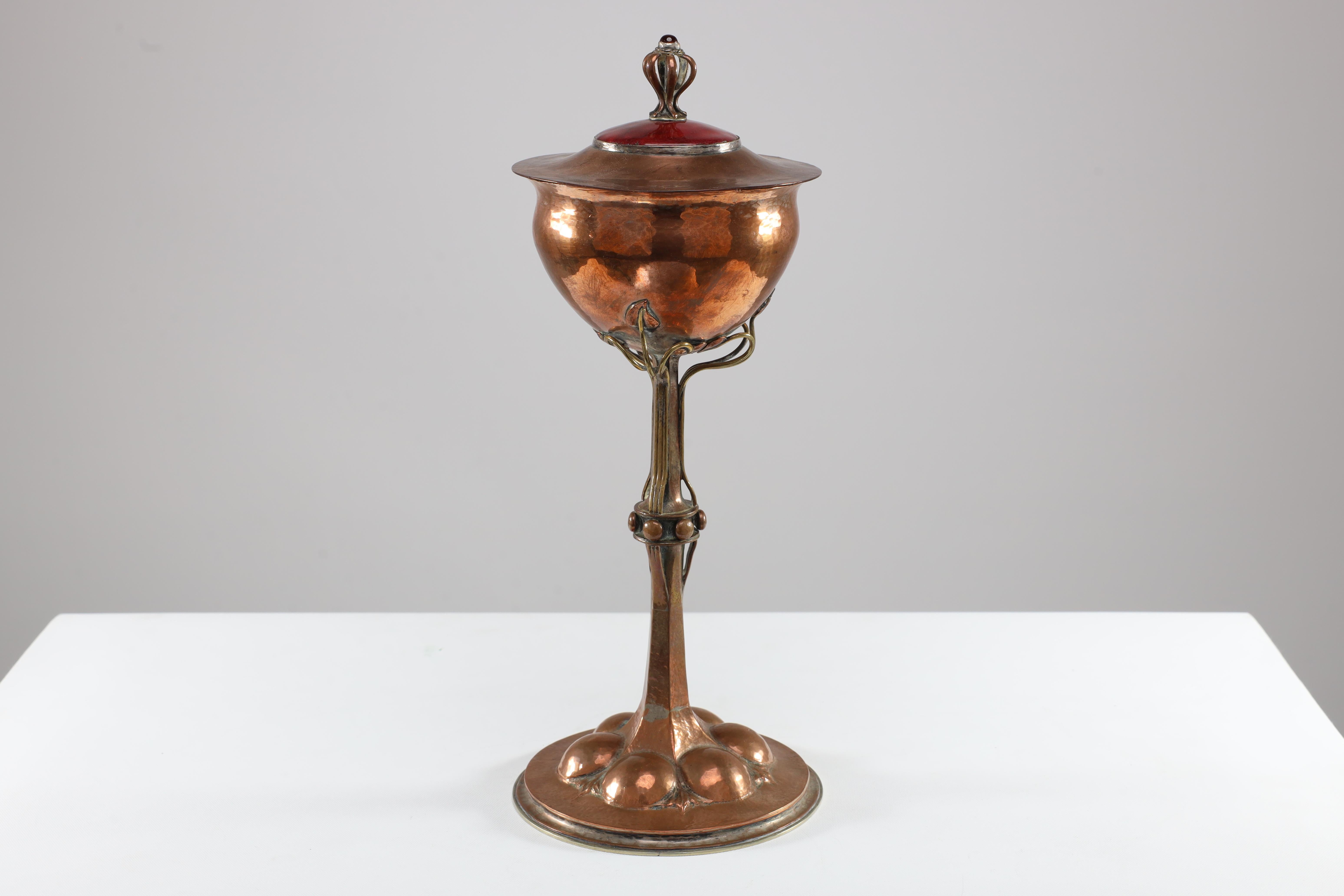 C R Ashbee. Made by The Guild of Handicrafts. An Arts and Crafts Copper chalice. The lid with semi-precious red stone to the wirework handle rising from a red enamel circular plaque. The bowl is attached to the stem with sinious wirework emulating