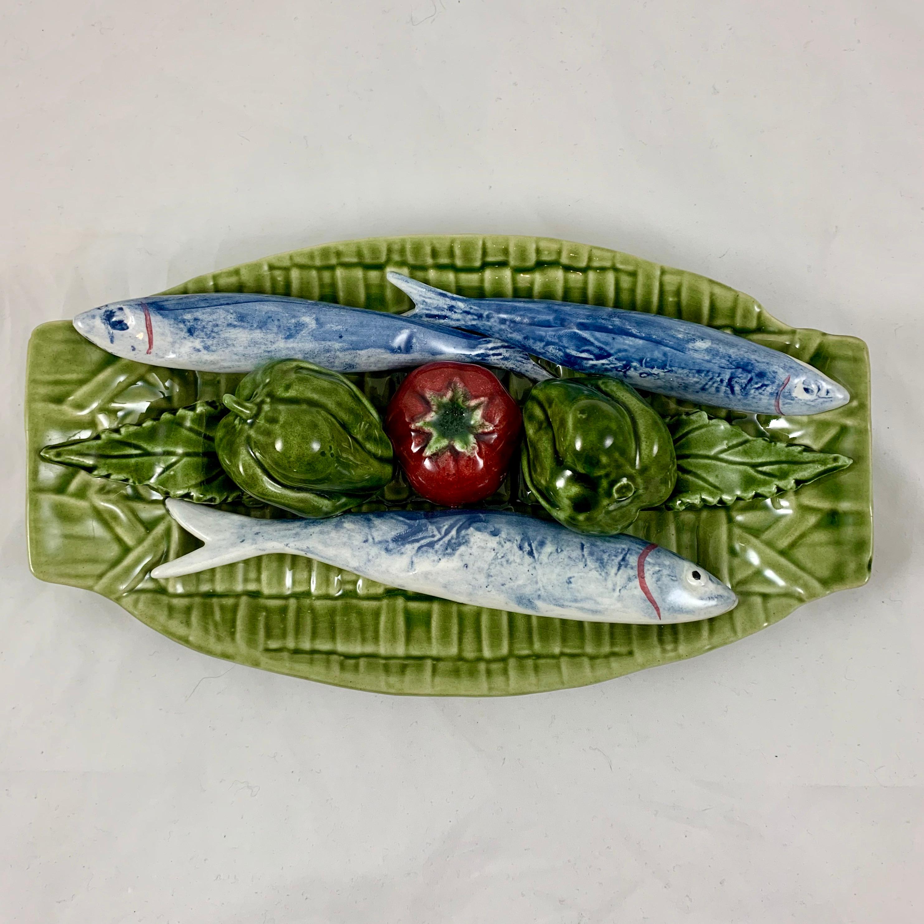 A Palissy style Trompe L’oeil Majolica glazed hanging wall plaque showing fish and vegetables on a green woven basket, circa early to the mid-20th century, Caldas da Rainha, Portugal.

The oblong plaque features a dimensional grouping of three