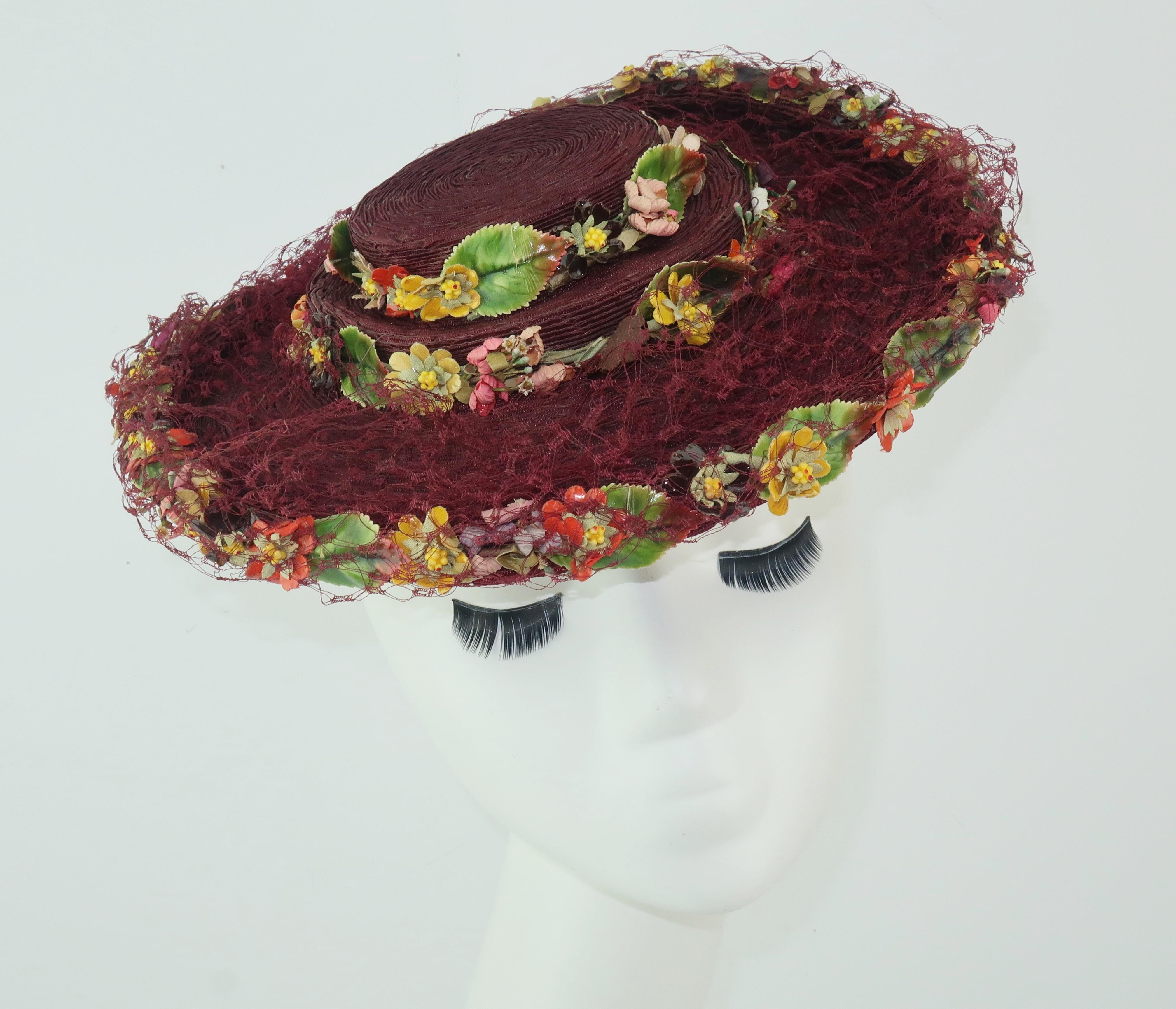 A C.1940 deep burgundy straw hat by Atlanta milliner, Ella Buchanan Gunn, embellished with lacquered silk flowers and netting.  The silk flowers introduce shades of green, coral red, yellow, aubergine and pink.  The unique shape of the hat is