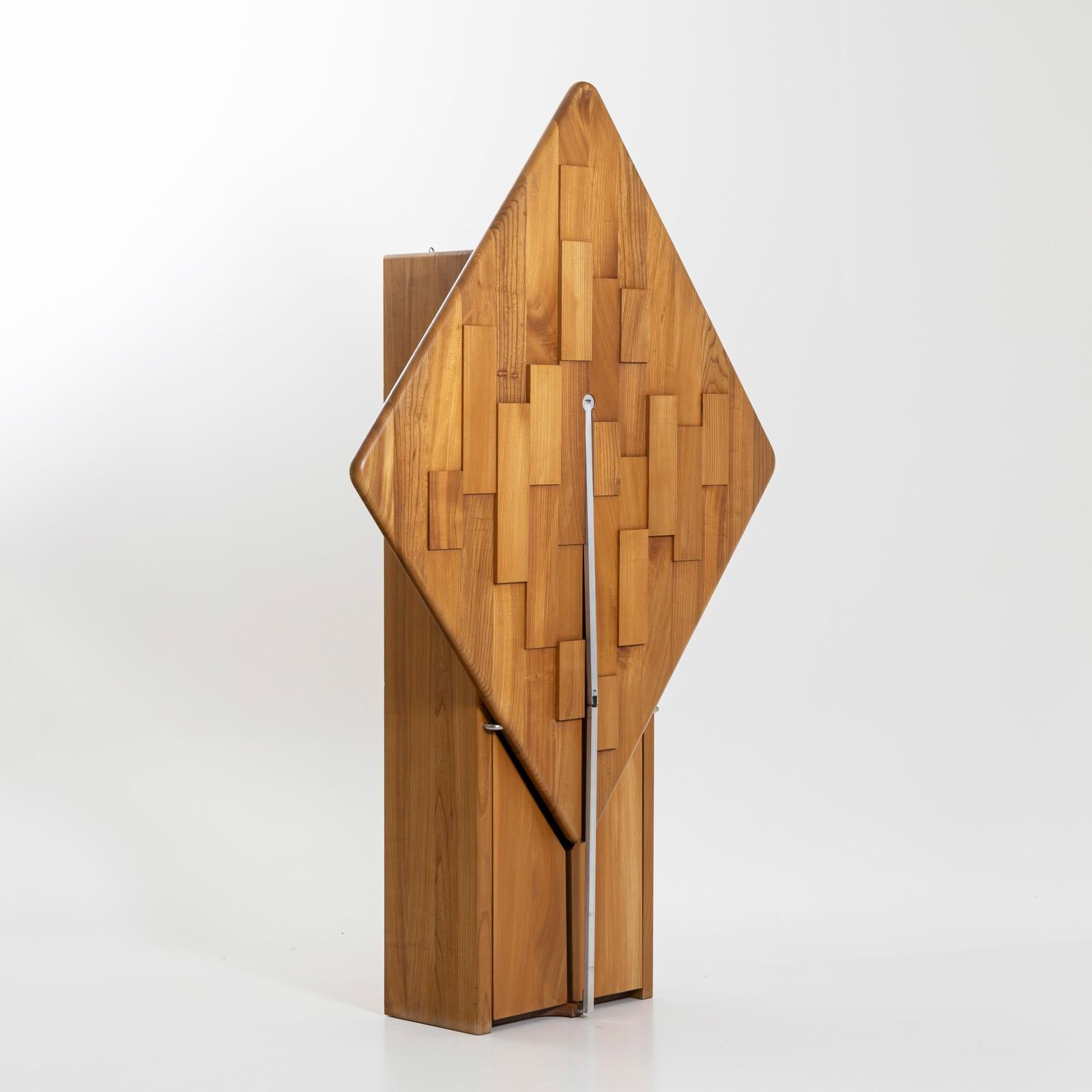 Narrow wooden cabinet with two doors and glass shelves in the lower section and a fold-down table top. The table top is diamond-shaped and decorated on the underside or outside with rectangular wooden discs in a geometric pattern at different