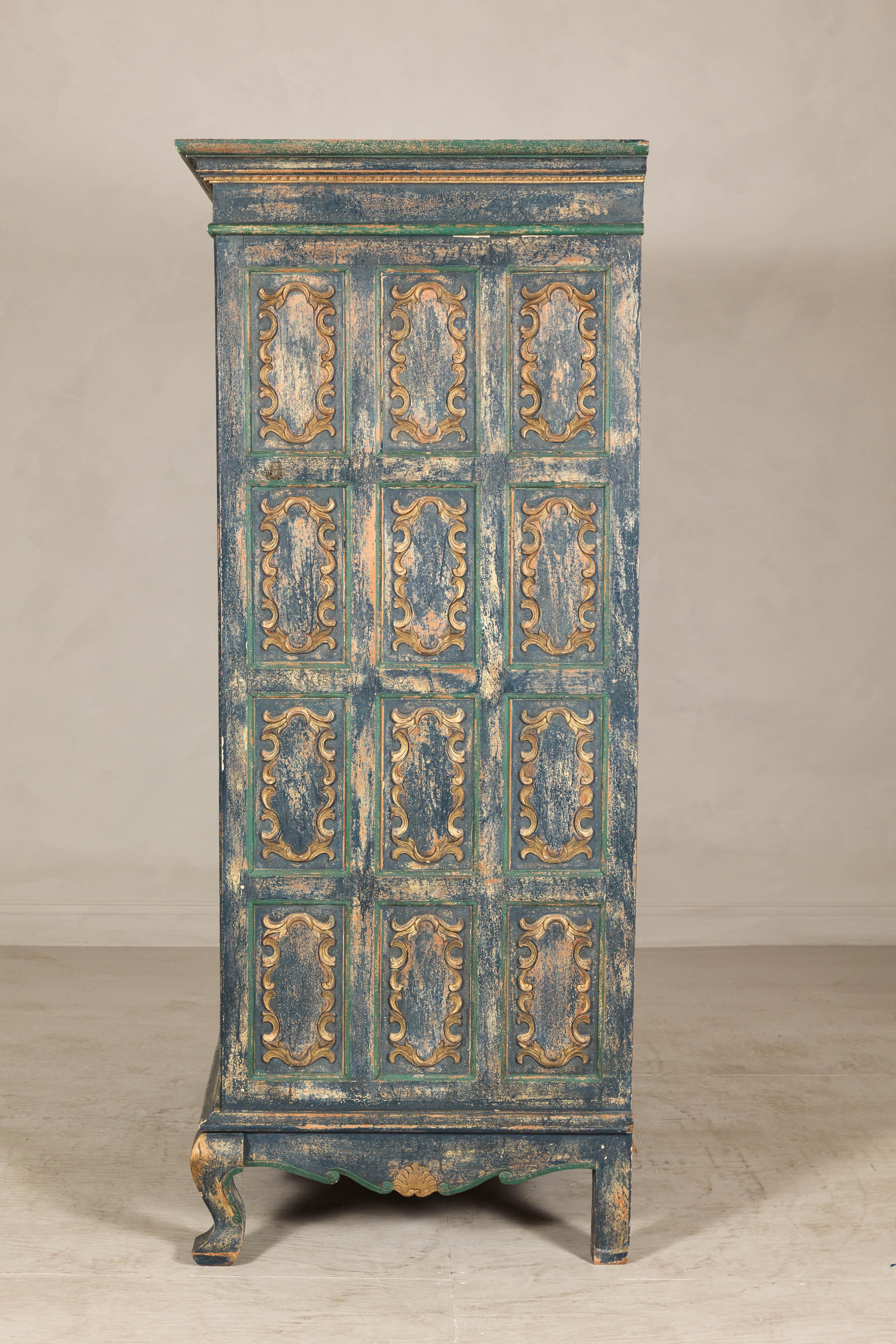 A Dutch Colonial cabinet with blue patina and cabriole Legs. This tall cabinet offers a powerful statement piece rooted in the decorative language of Dutch Colonial design. The facade is composed of deeply paneled doors, each inset with gilded