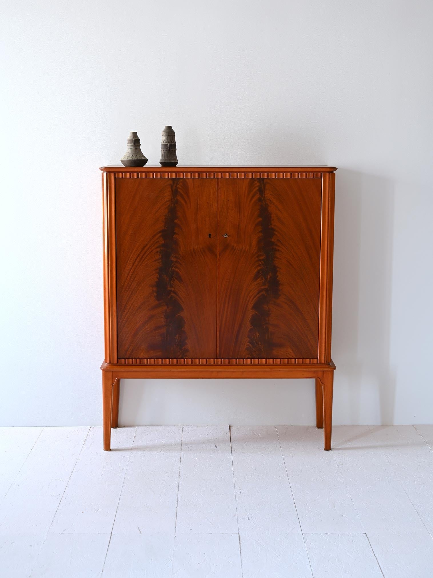 Cabinet vintage dal design raffinato con dettagli artigianali. Le ante a battenti in legno sono caratterizzate da marcate venature che formano un gioco geometrico di forme. Ai lati, il legno è intarsiato con listelli verticali. 

Le gambe lunghe e