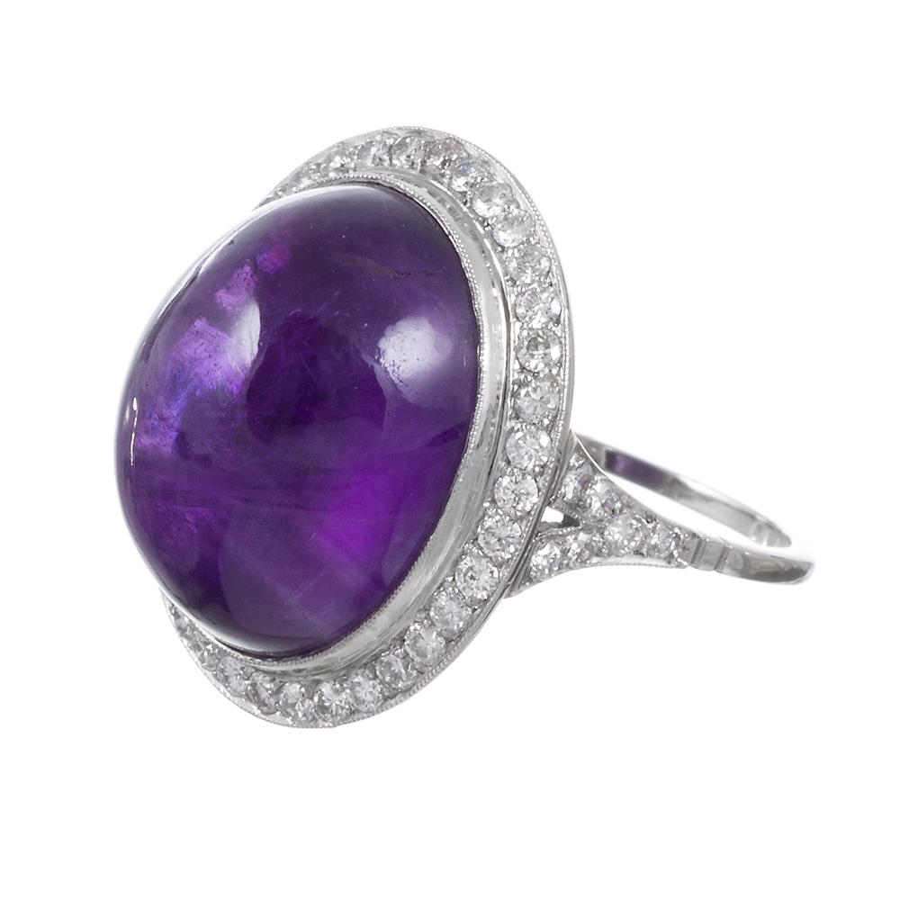 An alluring and beautifully detailed variation of a classic cluster style ring, rendered in platinum, with a large cabochon amethyst at the center of a frame of .75 carats of brilliant diamonds. The drama of a larger piece is tempered by the smooth