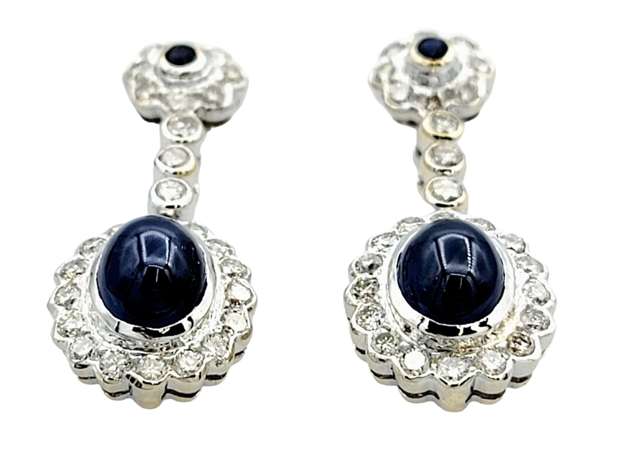 These exquisite dangle earrings, set in luxurious 18 karat white gold, are a harmonious blend of sophistication and intricate design. The focal point of each earring features an elegant oval cabochon sapphire, exuding a timeless allure and
