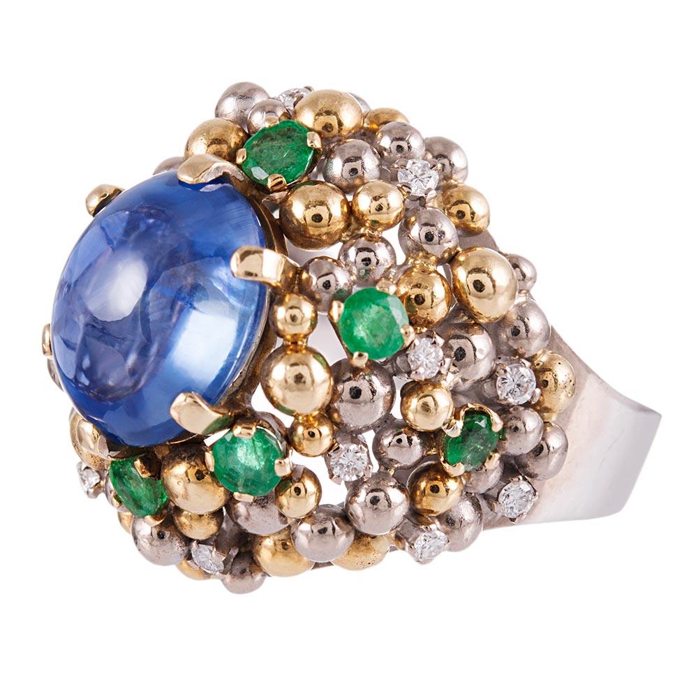 Made of 18k white and yellow gold, the 2.50 carat cabochon sapphire sits atop an artful cluster of faceted emeralds, golden orbs and brilliant diamonds peppered about the landscape. The emeralds and diamonds weigh .60 and .40 carats in total