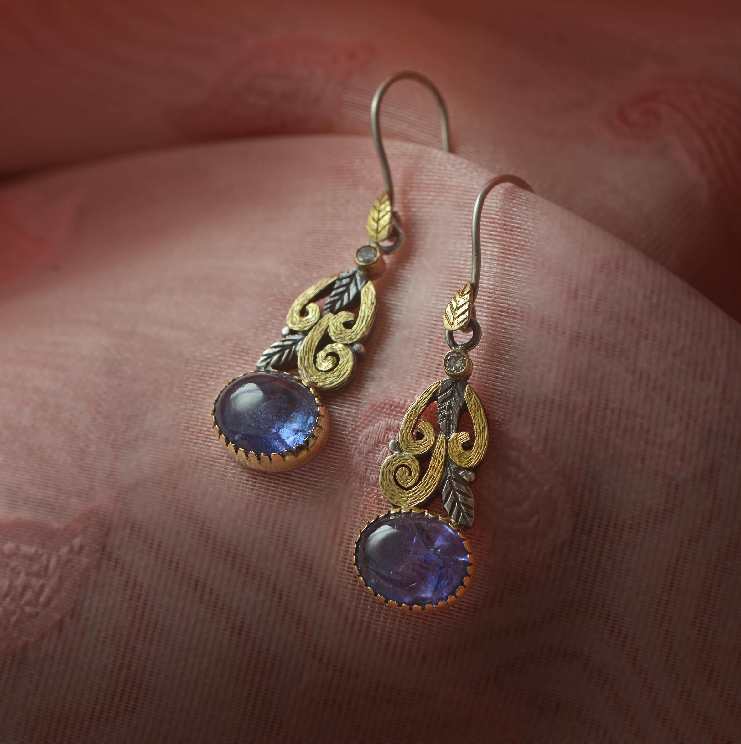 These one-of-a-kind dangle tanzanite earrings have been handmade in our workshops. They feature cabochon tanzanites which are capped with full cut diamonds and set in 18ct gold and oxidized sterling silver hand-engraved geometric and botanical