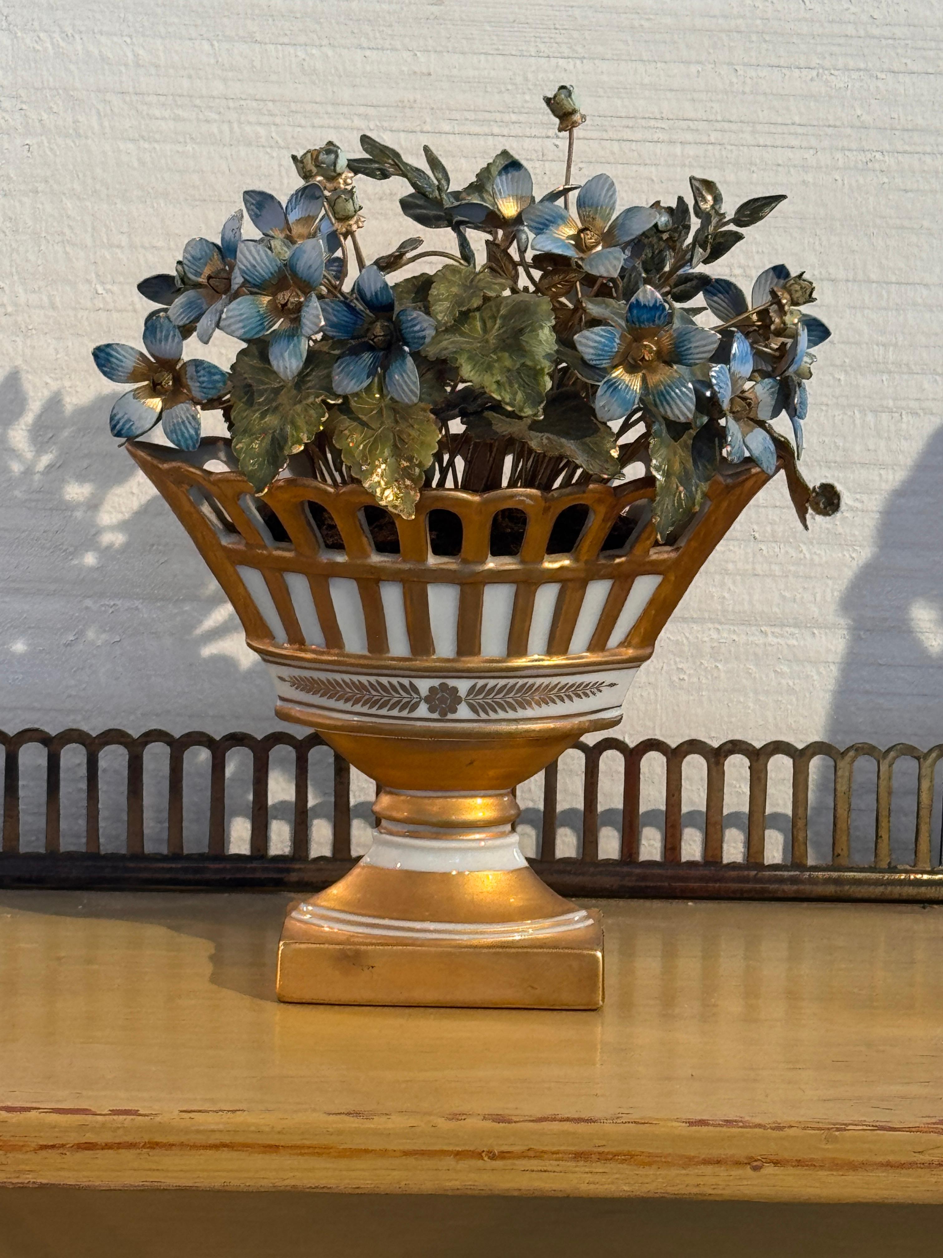 Petite composition de fleurs émaillées dans les tons bleus et verts, placée dans une urne en forme de panier, dorée et blanche, décorée d'une bande florale. La pièce reflète le travail finement détaillé de Hutchison avec des pétales et des feuilles
