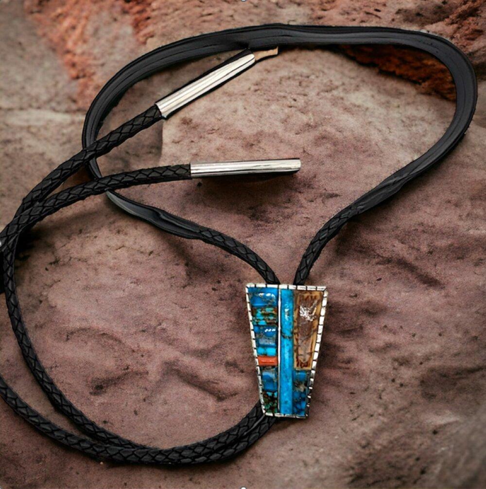 This hand-fabricated bolo tie by Rob Sherman, presented by Cactus Flats, is a true masterpiece of Southwestern jewelry. Featuring hand-cut Kingman turquoise, coral, and mammoth bark, expertly arranged to create a unique and eye-catching design, this