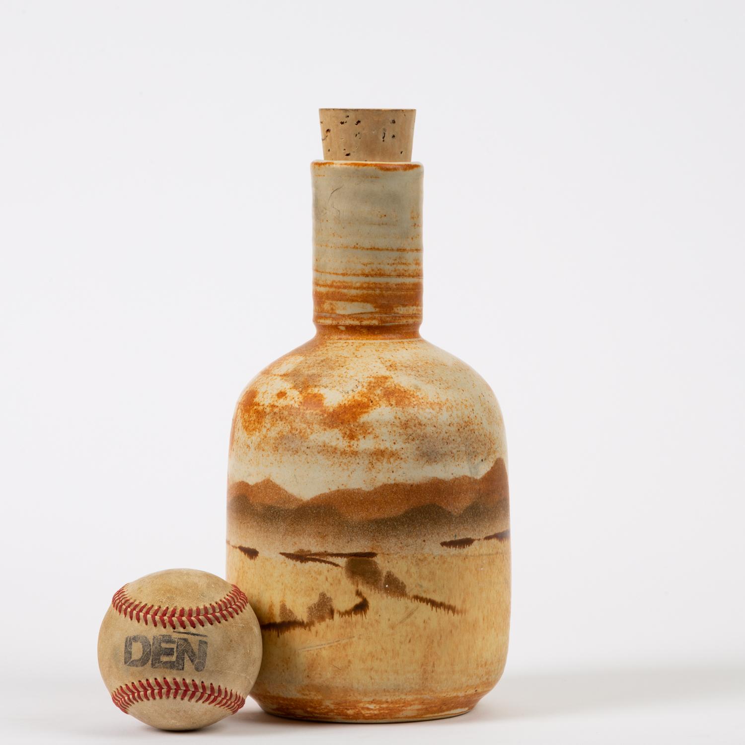 A studio pottery ceramic bottle with a cork stopper. The wheel thrown piece has a wide body with sloping shoulders, tapering towards the foot, and is notched where the neck joins the body. The vessel features an earth-toned color palette in tan,