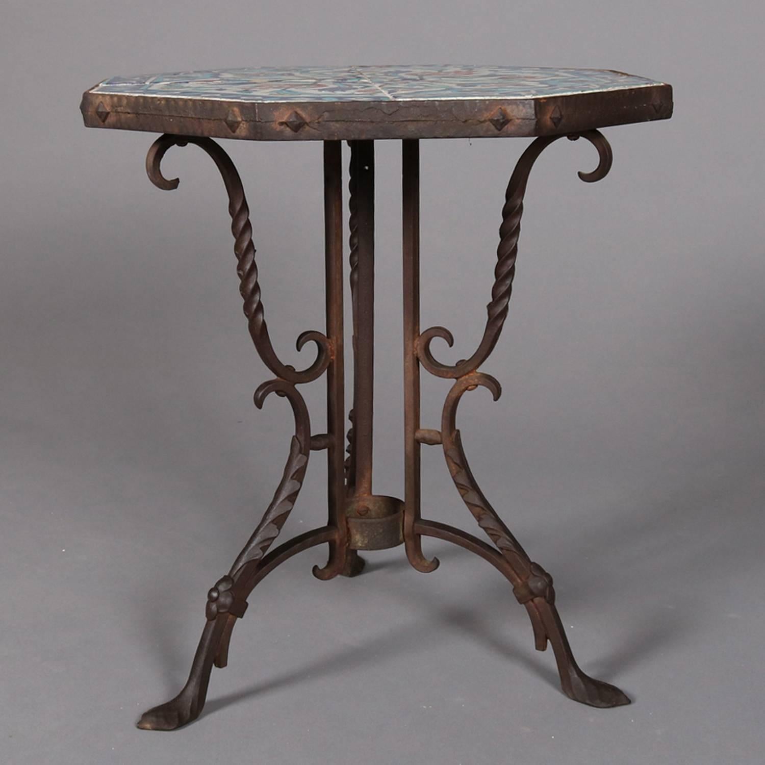 California Yellin School plant stand features wrought iron base with tripod twist foliate and scroll form legs supporting octagonal mosaic tile top with urn and floral decoration primarily in blues and seated on snake head form feet, circa