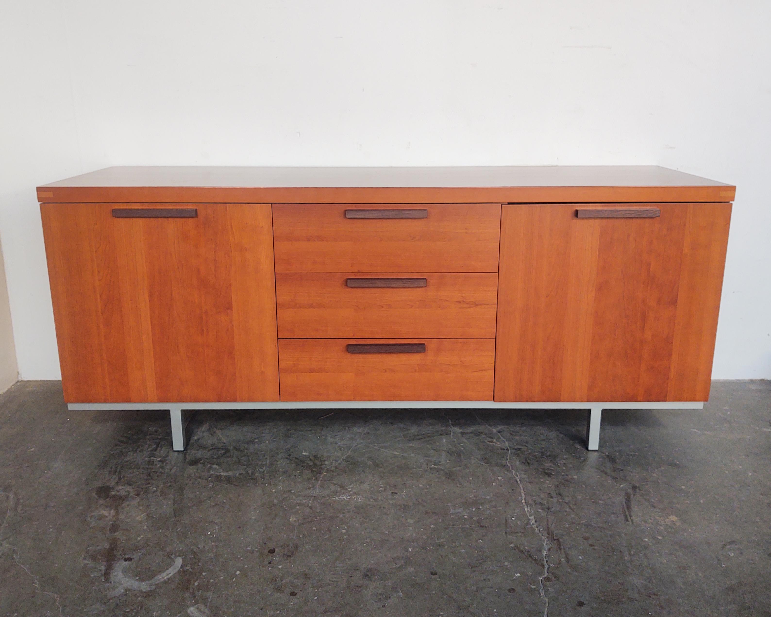 Cherry wood credenza by Calligaris, made in Italy. Minimal design with metal base and exposed top corner joinery. Three drawers down the center with solid oak ebony-stained wood pulls. Cabinets on each side feature adjustable glass shelf. Overall