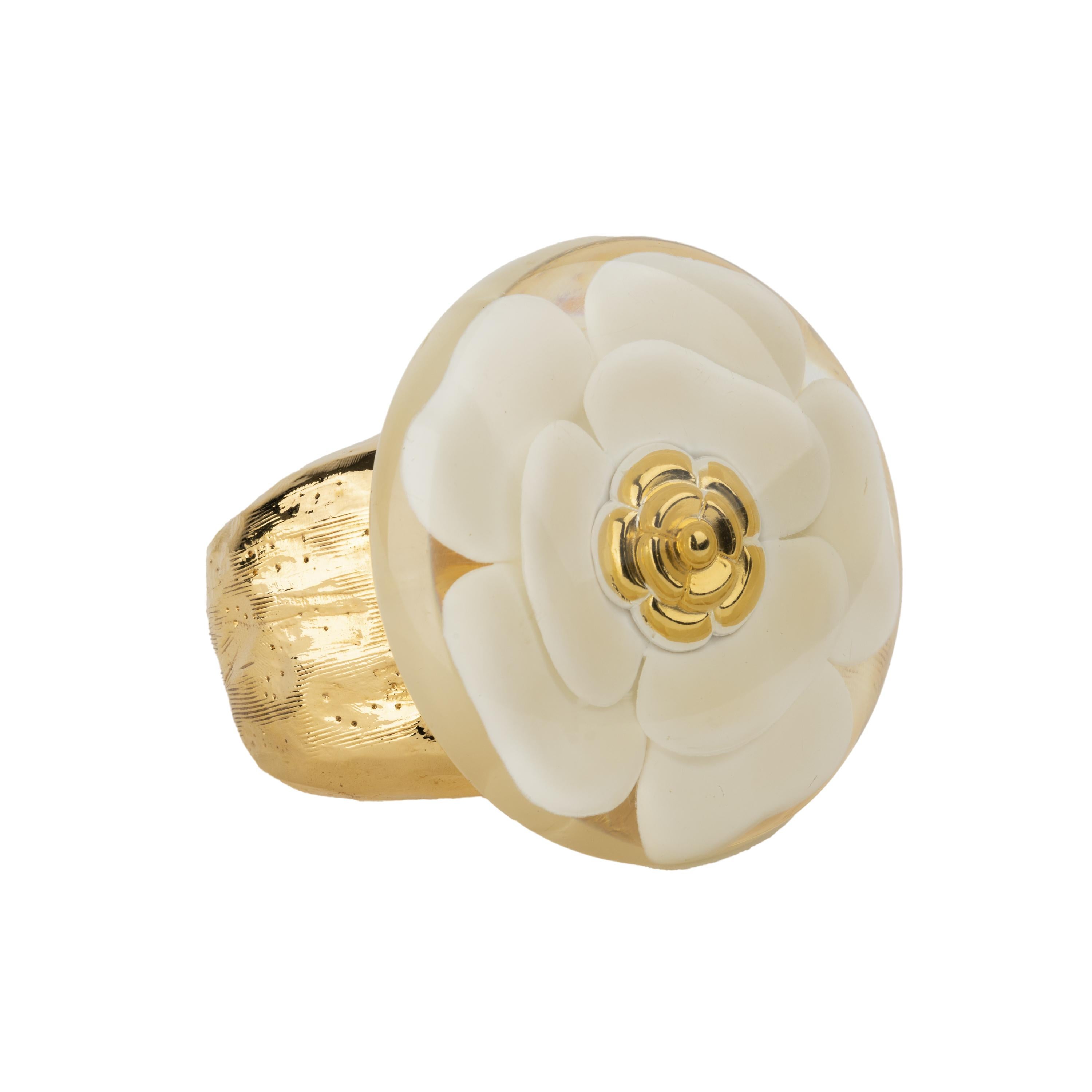 Bracciale Chanel in metallo placcato oro. Stile rigido aperto, decorato con il simbolo della famosa casa di moda: la camelia bianca, posta all'interno di un disco in acrilico lucido trasparente.

È disponibile l'opzione NFT.