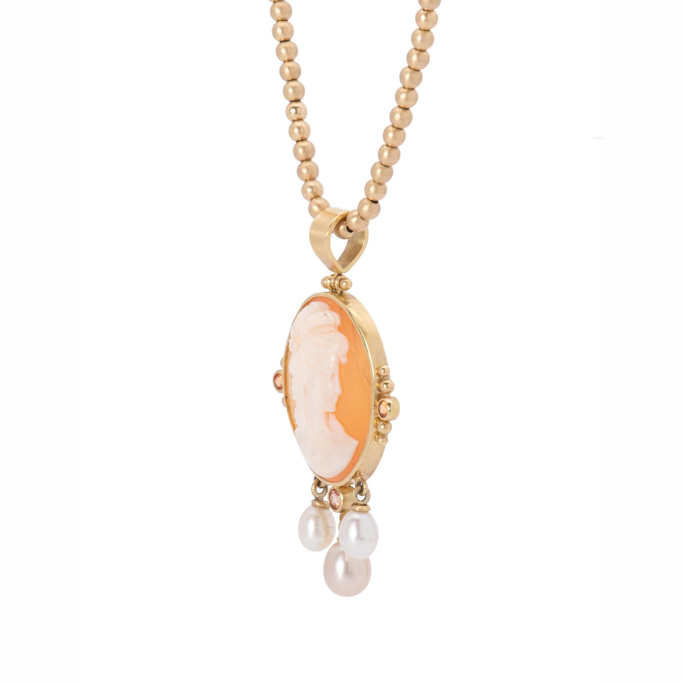 A classic carved shell cameo depicting the profile of a Regency-era lady is bezeled in 18k gold and fitted with gold beads, orange sapphires .28ctw and a generous hinged bail. The cameo and pearl pendant in 18k gold is embellished with a trio of