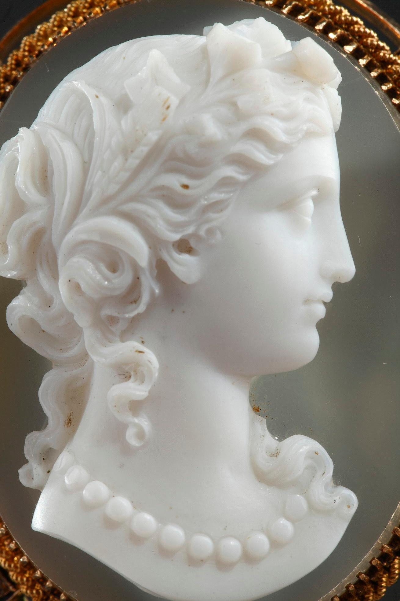 Cameo on blue-gray agate featuring a young woman looking toward the right. The artist intricately sculpted the white vein of the agate to bring the delicate profile to life. The young woman represented in bust has her hair raised and adorned with a