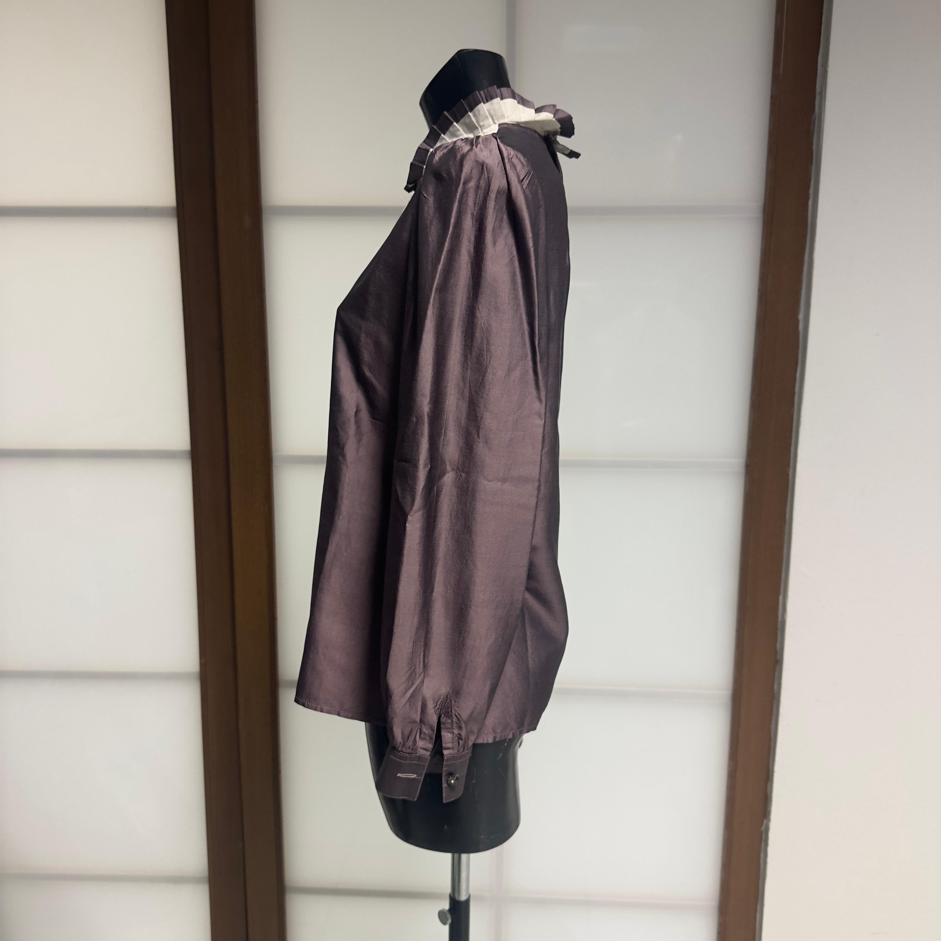 Questa elegante camicia Prada, realizzata in Italia nei primi anni 2000, rappresenta perfettamente l’estetica essenziale e intellettuale della Maison.
Il modello è costruito in un raffinato taffetà dall’effetto luminoso color malva, abbinato a un