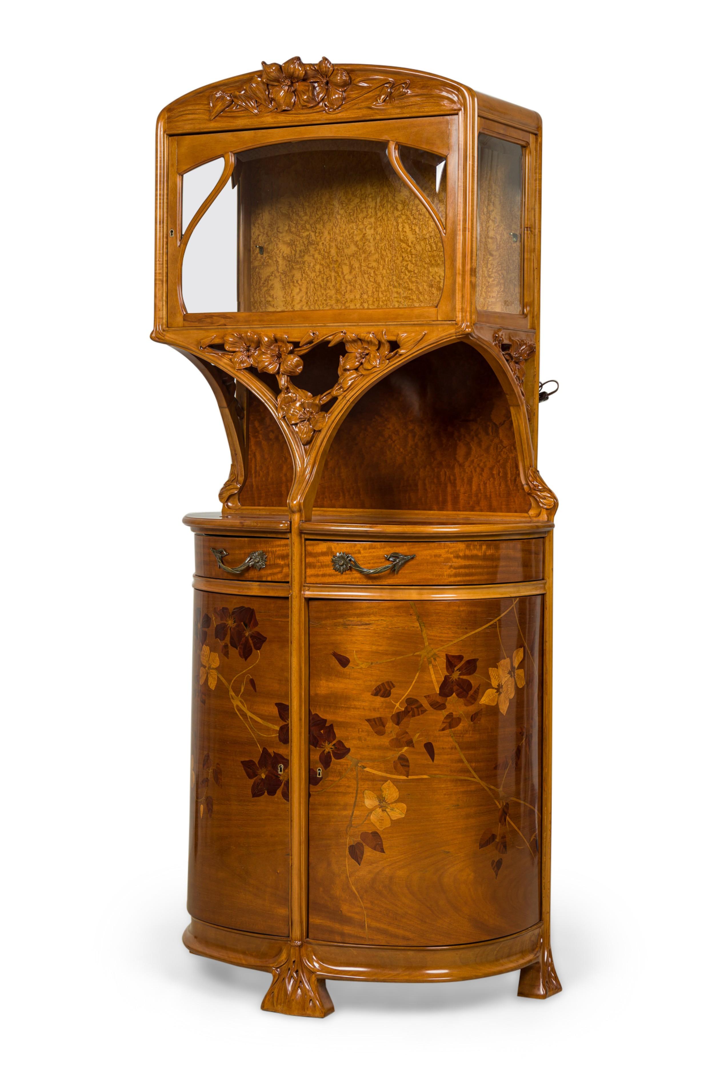 French Art Nouveau two-tier display cabinet / vitrine with a glass upper cabinet topped with an elaborately carved floral crest over an open compartment, resting on a lower demilune closed cabinet featuring two upper pivoting drawers with bronze