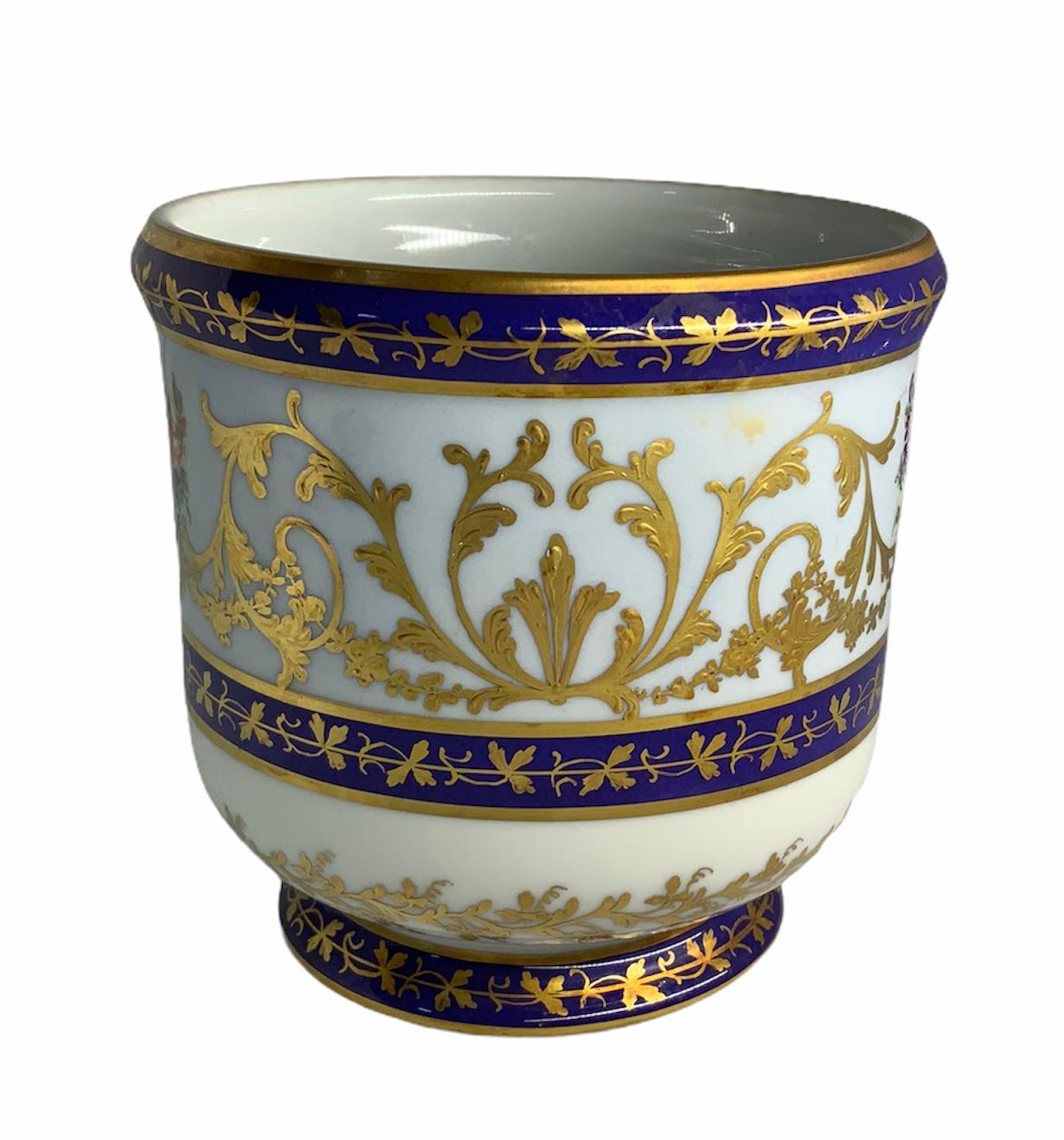 This is a hand painted Porcelain adorned with a gilded centerpiece vase with an arrangement of flowers in the center and gilt acanthus leaves growing around the sides of it. There are three cobalt blue rings around the cachepot decorated with a row