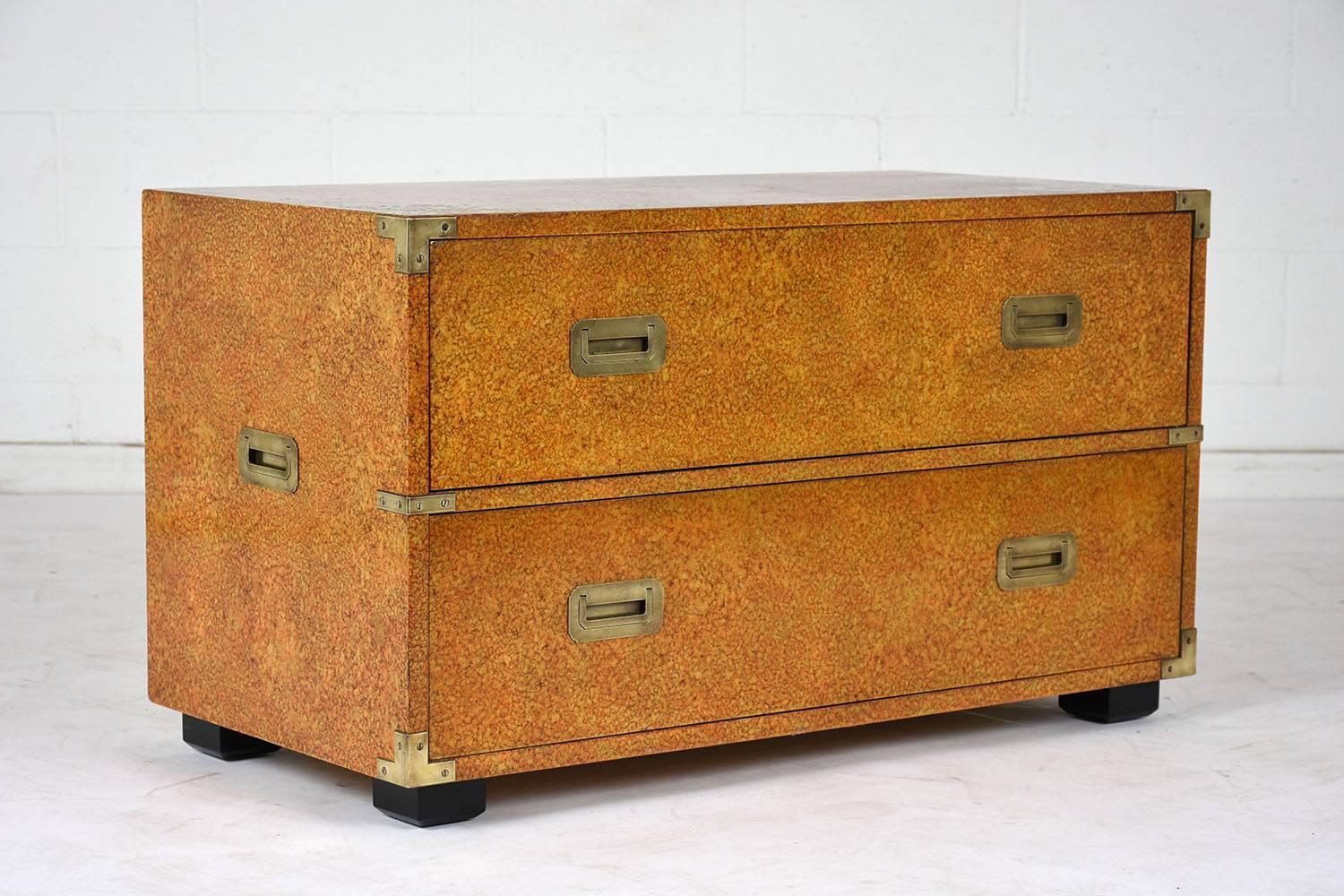 This 1970s campaign style chest of drawers is made by Henredon and features wood stained in a natural wood color with a sponge technique. The low chest has two drawers with flush brass drawer pulls. The chest has brass corner accents and brass