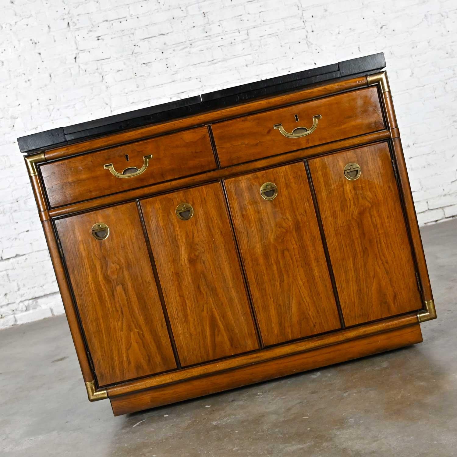 Fabulous vintage Campaign style dry bar in a medium-dark veneer, possibly pecan, with a black dyed wood top, an interior black laminate flip top, and antiqued brass plated hardware and casters by Drexel. Beautiful condition, keeping in mind that