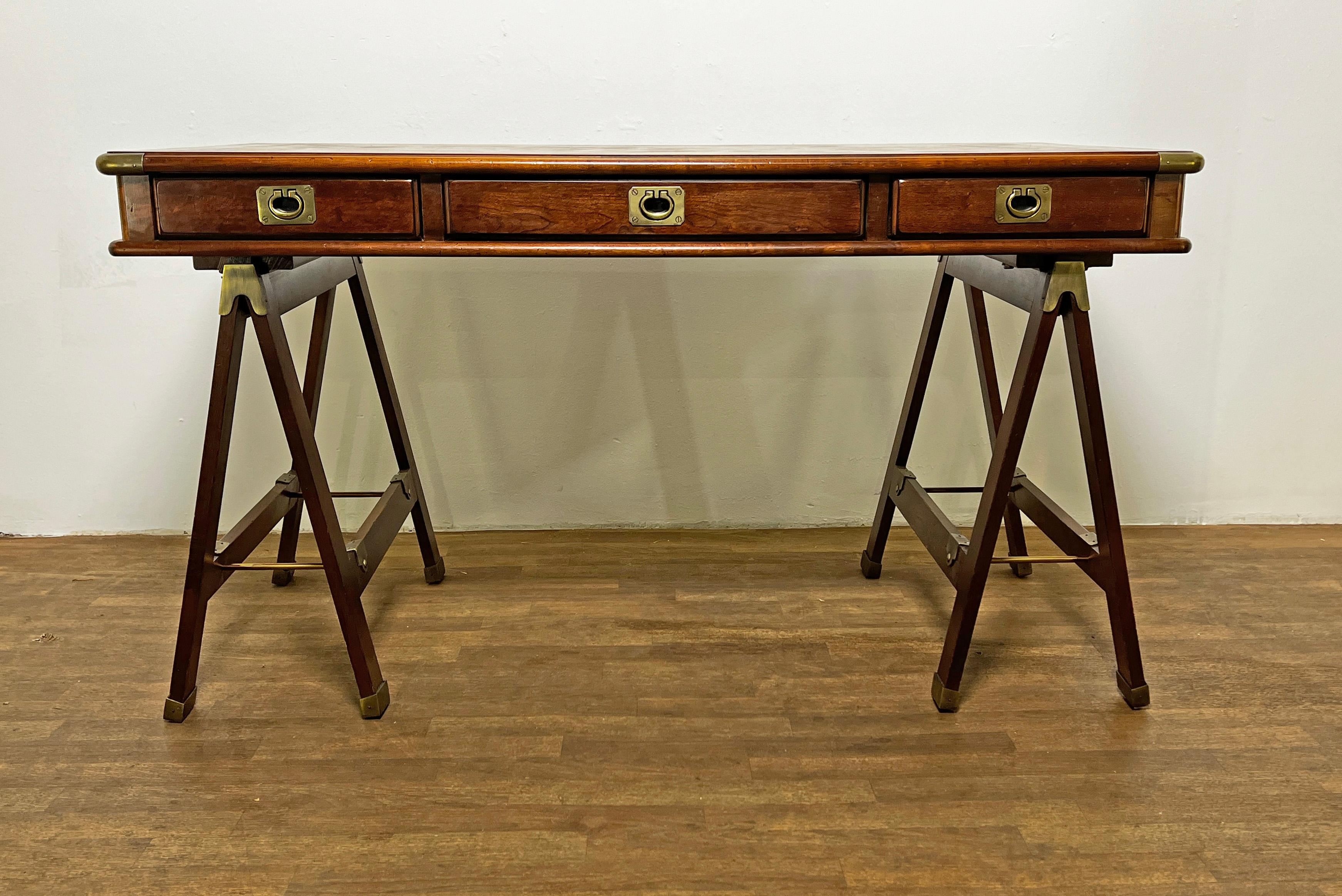 A mahogany campaign desk of three drawers, ca. 1970s, with brass bound corners and flush mount pulls on sawhorse legs 