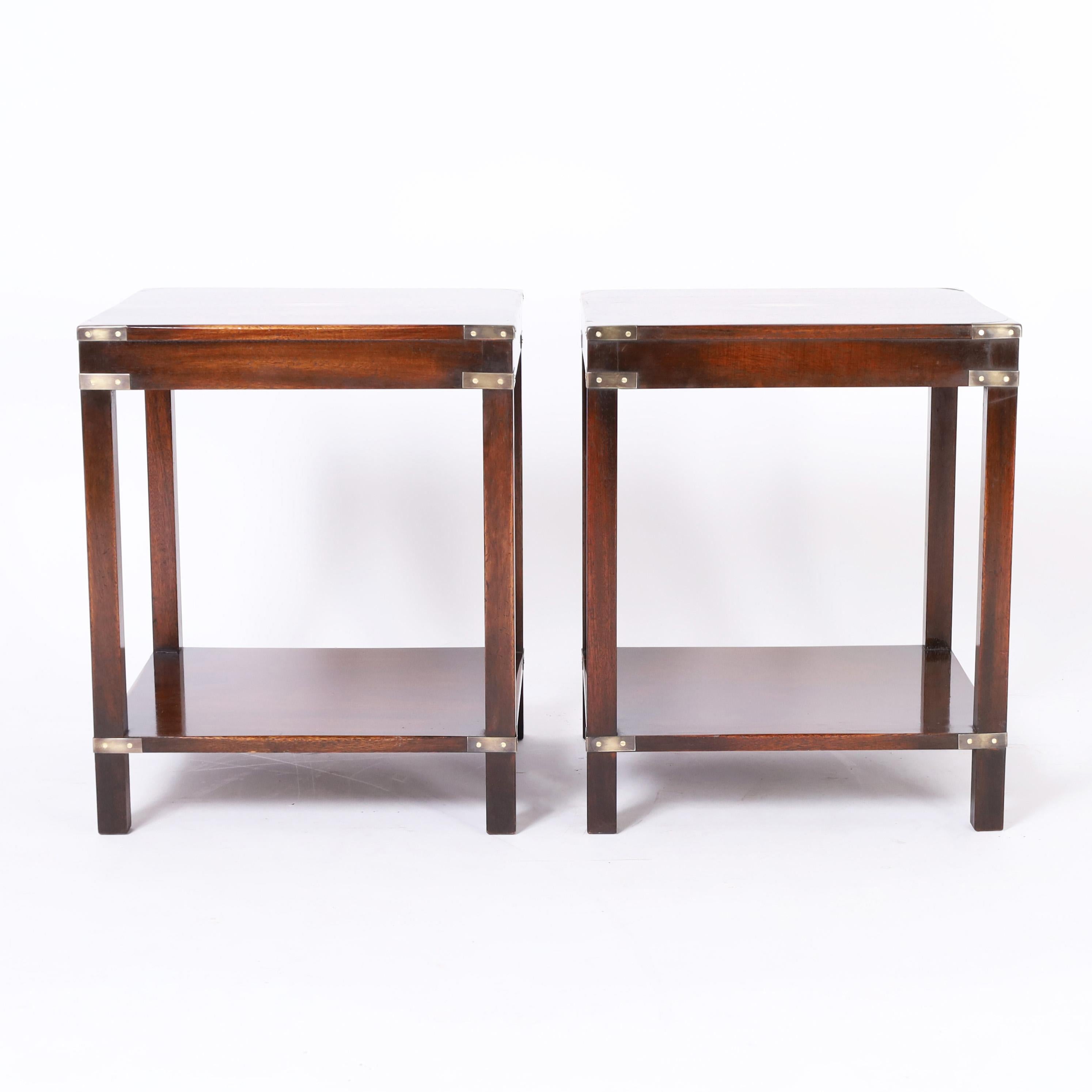 Handsome pair of British colonial style stands crafted in mahogany with two tiers in a stylized parsons form with campaign style brass hardware.