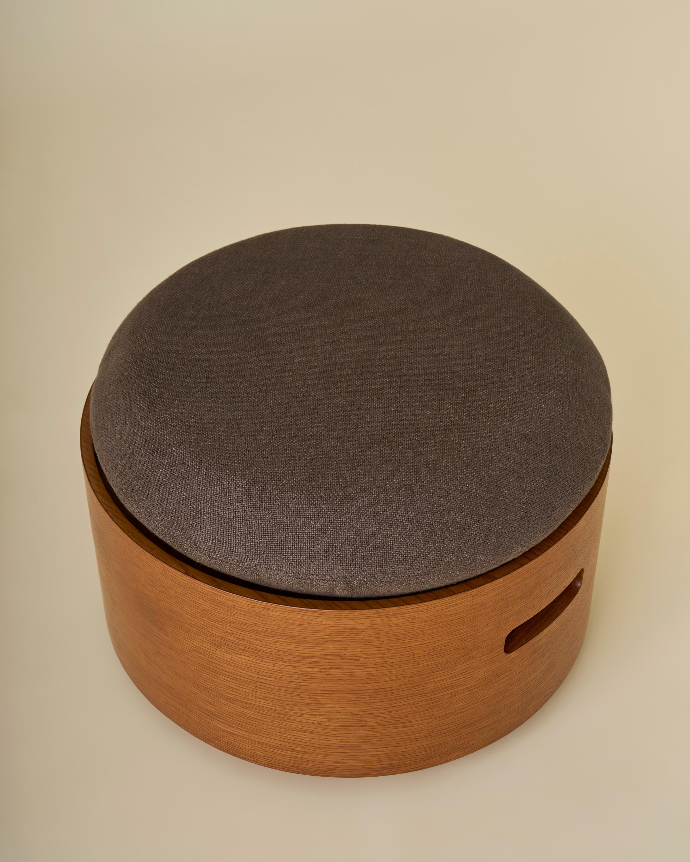 Le pouf Campbell est doté d'un plateau rembourré et de deux poignées de chaque côté de la base en bois de chêne.  Un subtil assemblage à queue d'aronde court verticalement sur un côté de la base, et la pièce dispose d'un rangement caché à