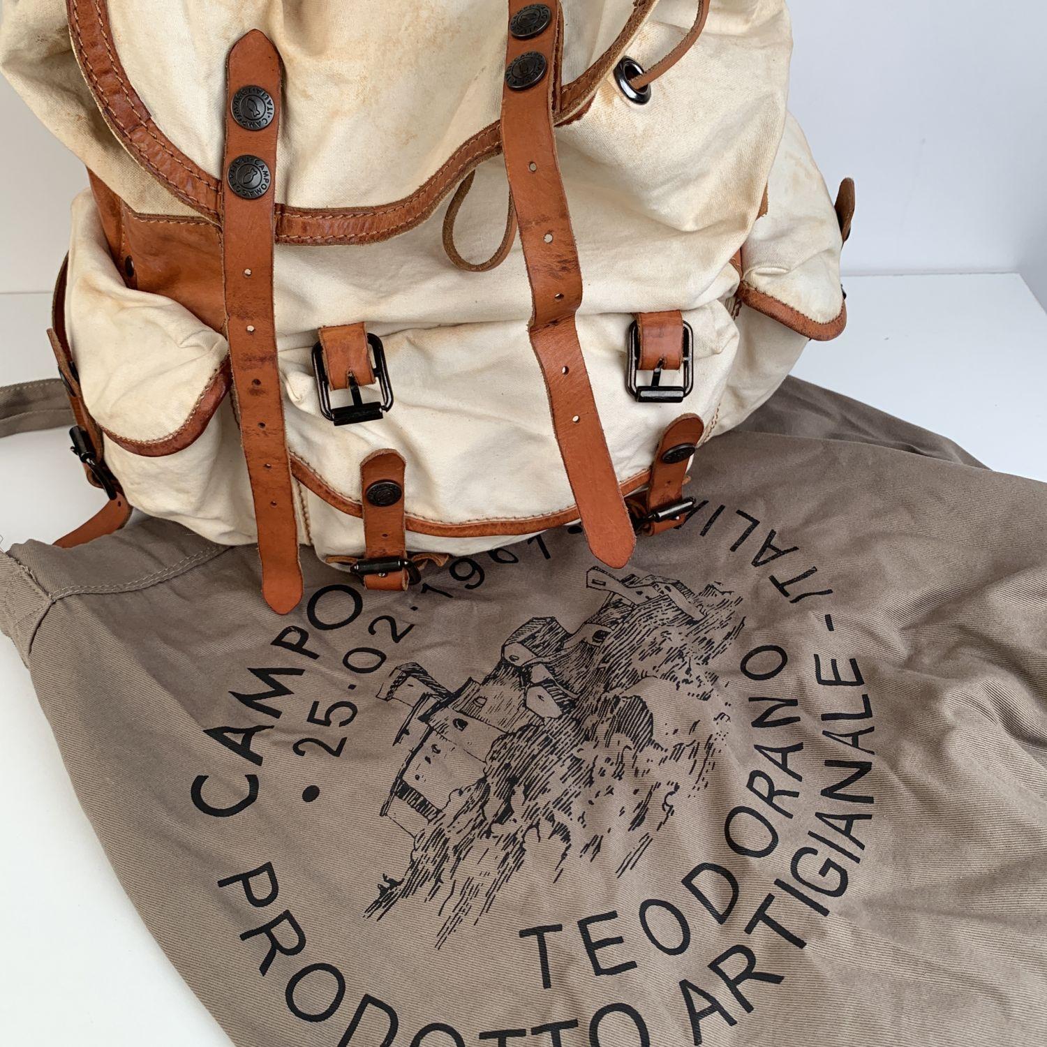 Campomaggi military style backpack crafted in ivory cotton canvas and trimmed with tan leather. Flap with double buckle/snap straps on the front + drawstring closure on top. Snap/buckle flap pocket on the front and snap/buckle flap pockets on the