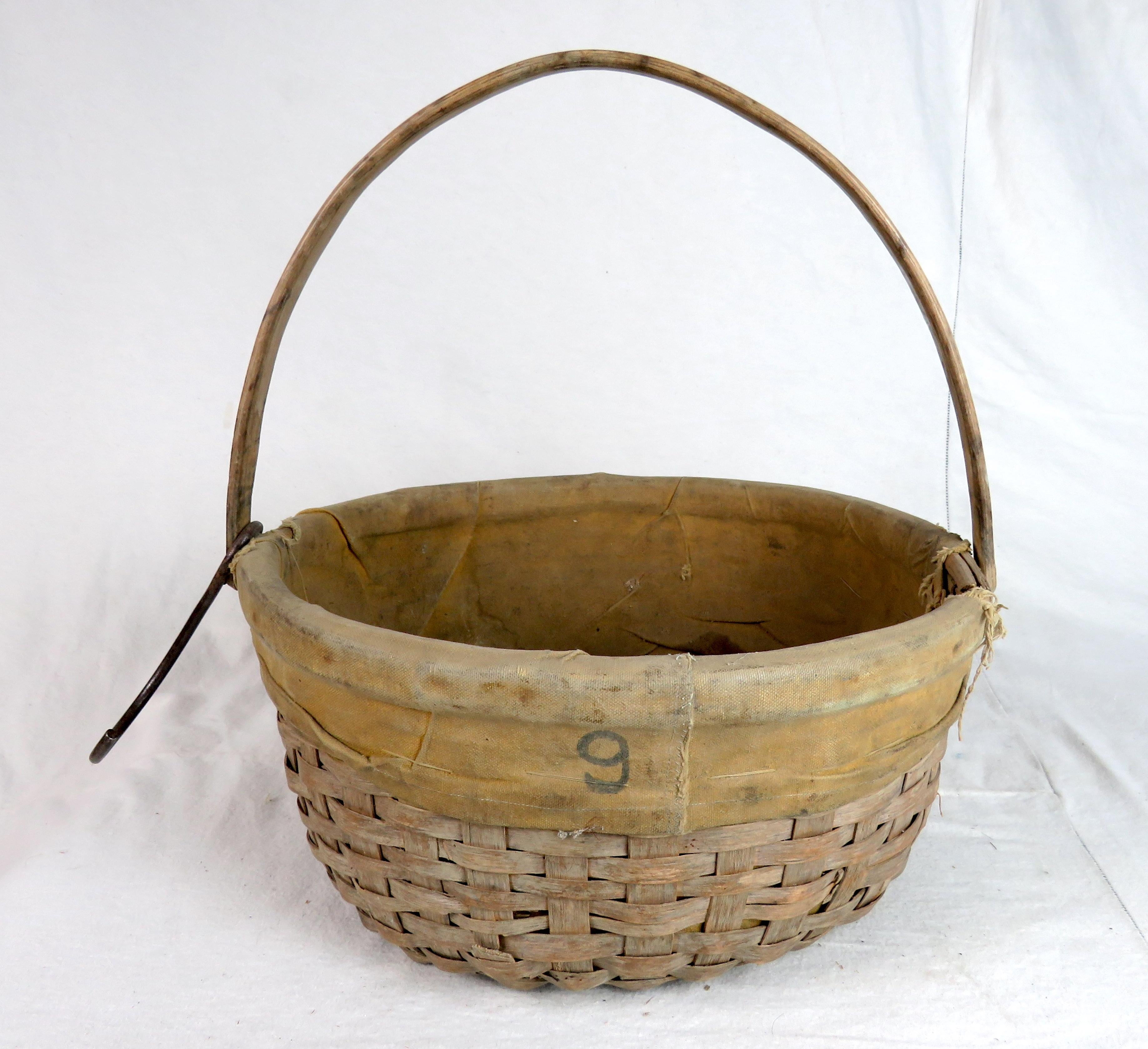 Turn of the century splint woven basket with swing handle, yellow canvas liner, and old hanging hook attached to its handle. Canvas with handwritten number 9 on one side and fraying at both handle connections from use.