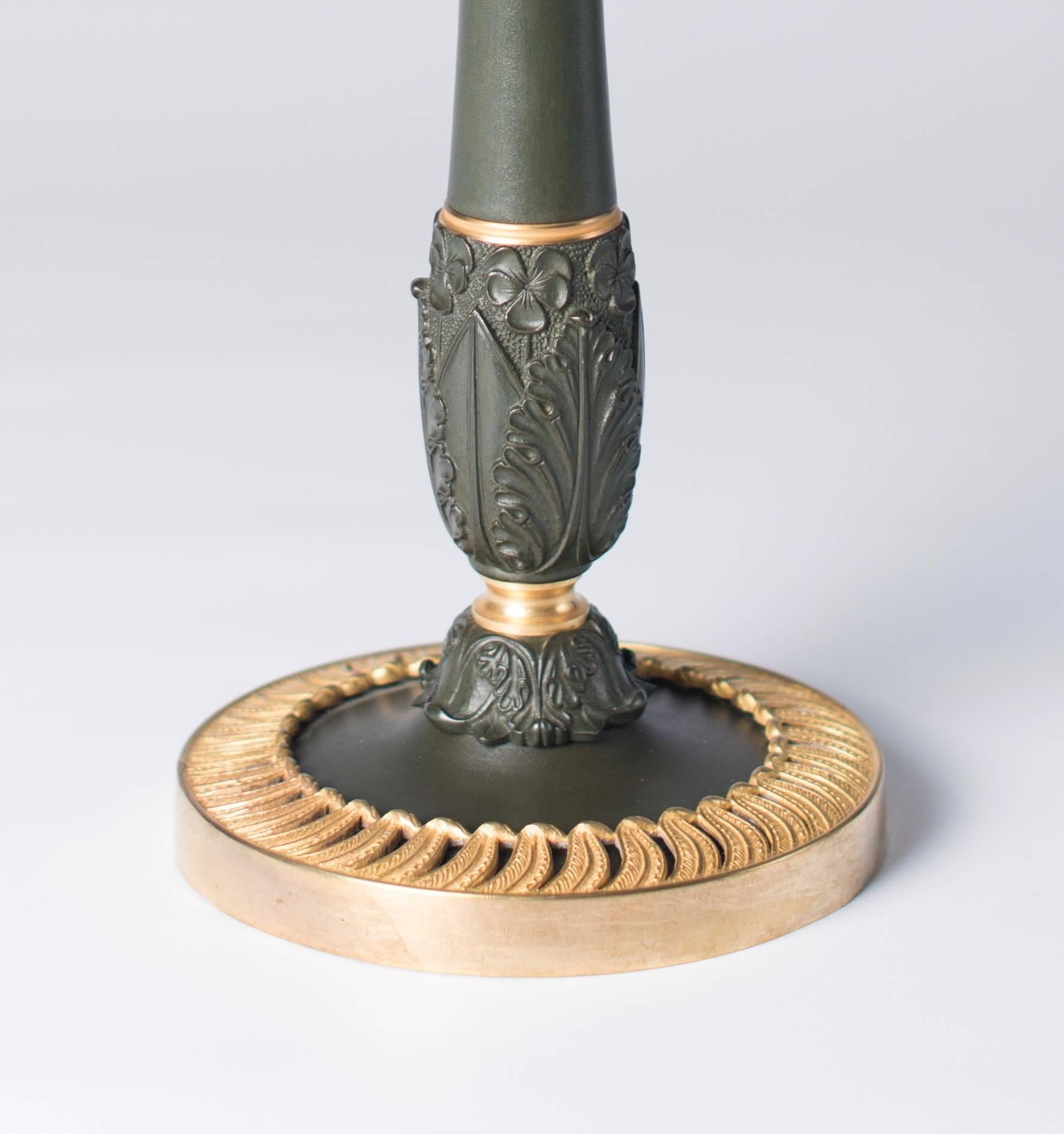 This pair of French Charles X gilt and dark olive green patinated bronze candelsticks do have a finely finished decoration. Each on a circular base with applied swirling open-worked band, above a baluster knopped stem and on top a vase-shaped