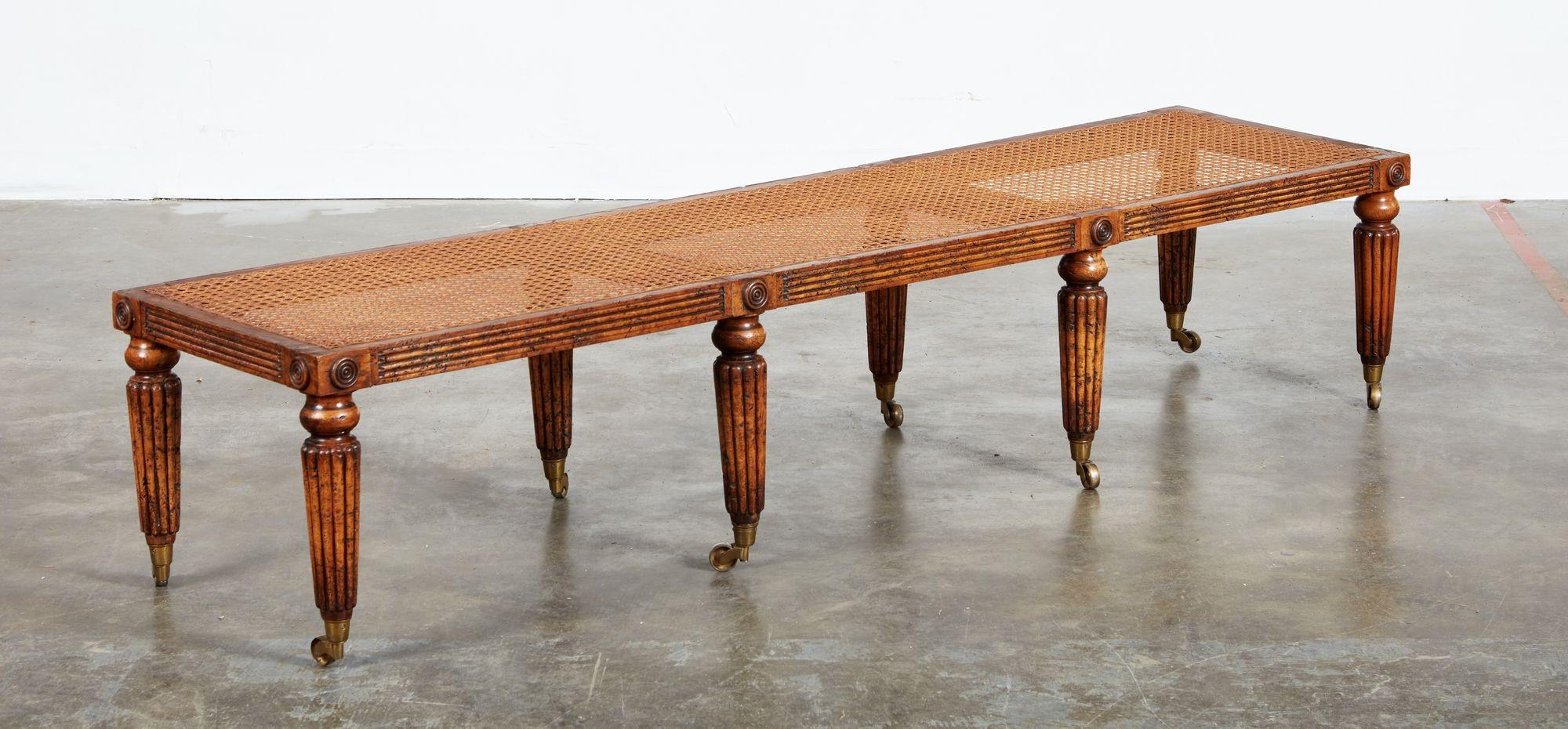 Un long banc plat en chêne têtard joliment marbré avec un siège canné au-dessus d'un tablier cannelé avec des rondeurs sur huit pieds cannelés terminés par des roulettes en laiton. Coussin d'origine fourni (montré sur les photos), et maintenant