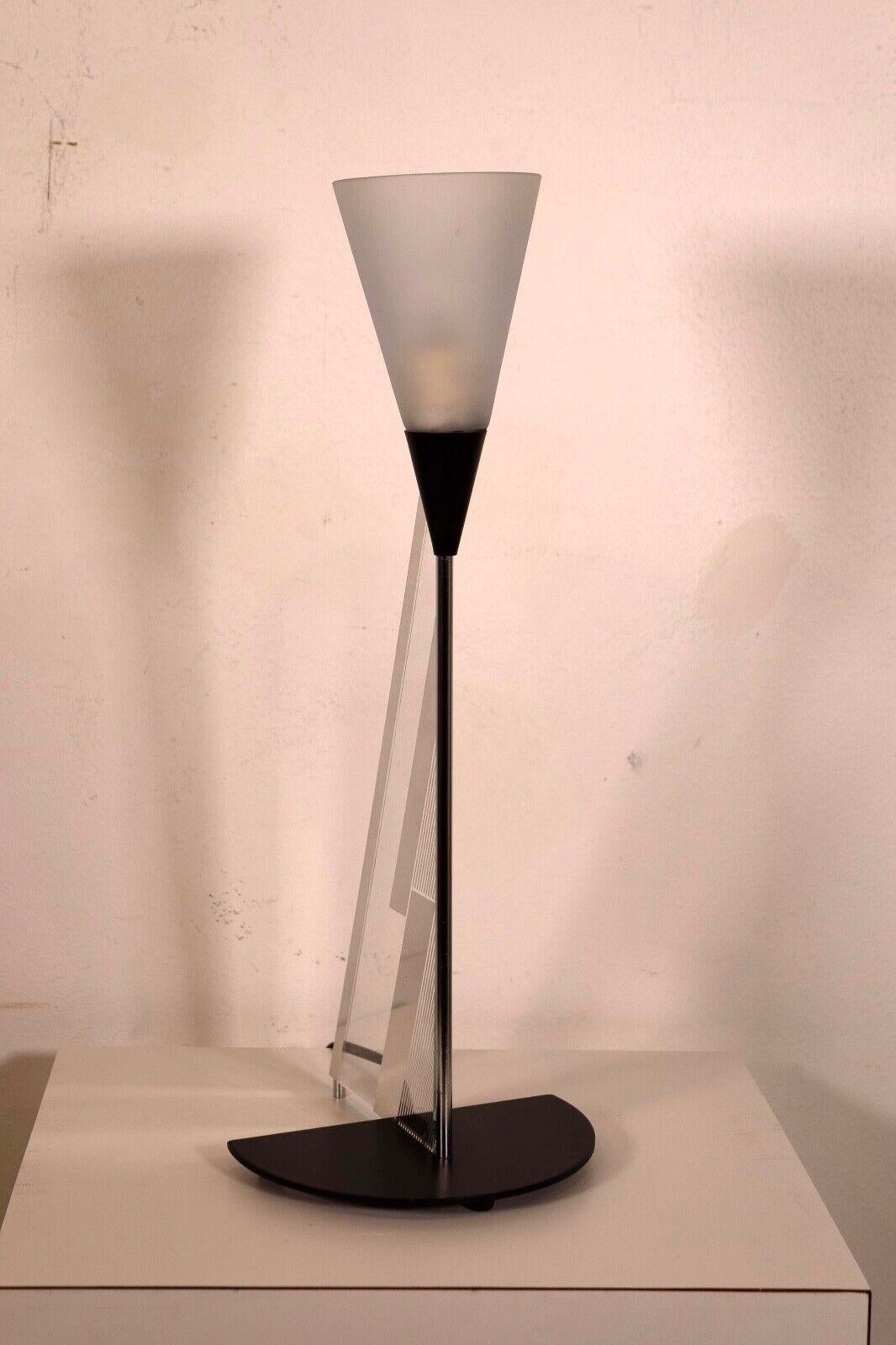 The Canetti Mid Century Modern Triangular Shaped Lamp features a sleek, geometric design with a sharp triangular shape that tapers towards the top. Its base is a black semi-circle that complements the lamp's modern aesthetic, providing a stable