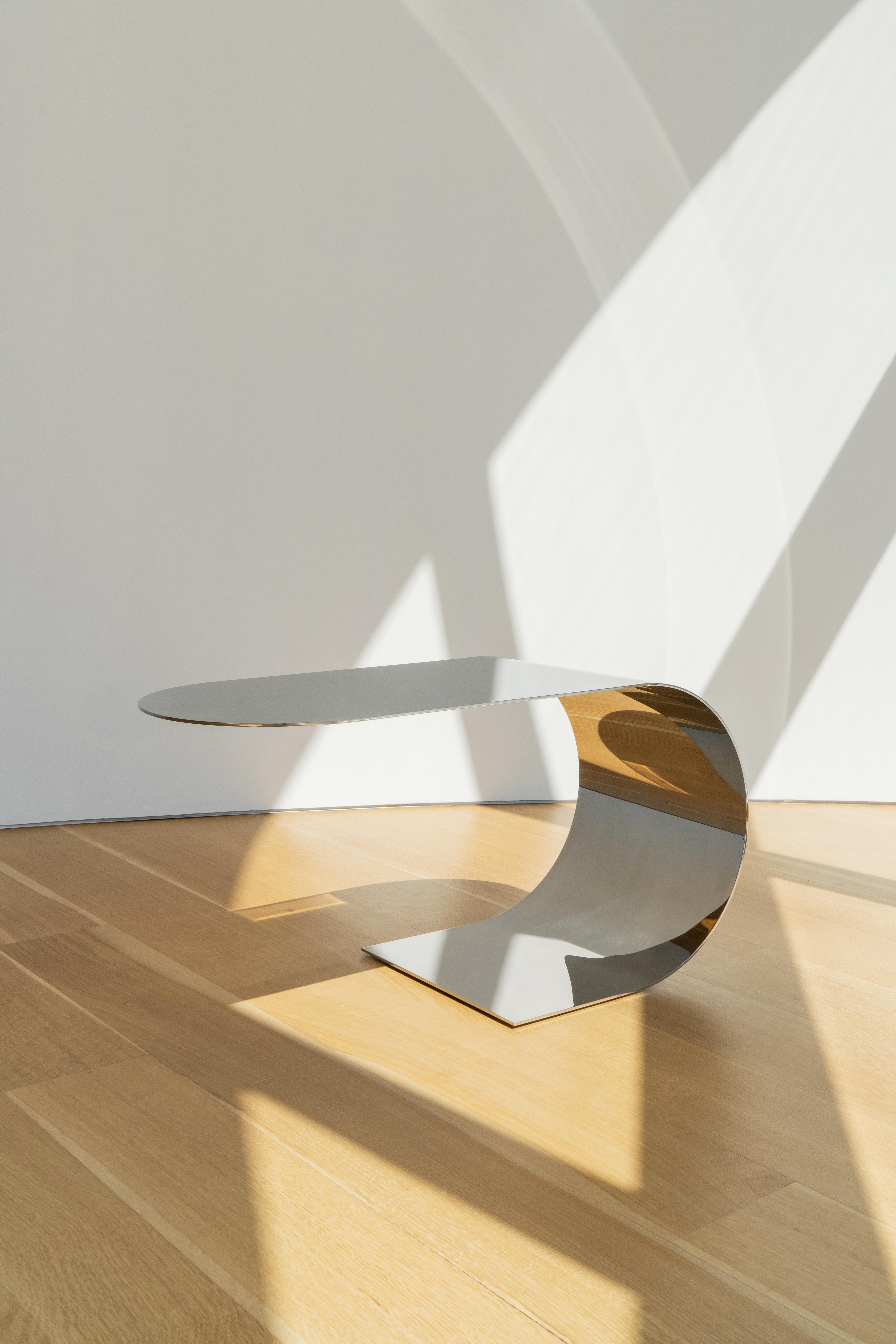 This extended c-shaped side table with cantilevered top is made of 3/16” recycled polished stainless steel or brass. It is also available in 5/8
