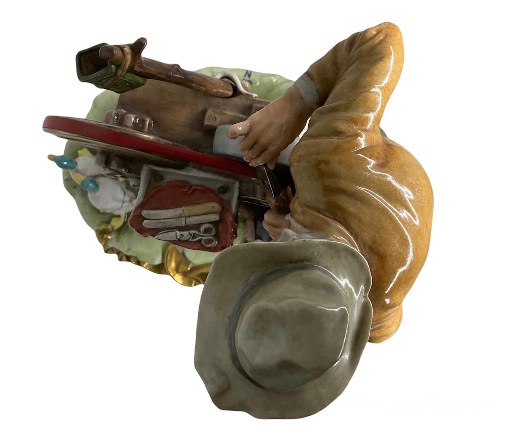 This is a large heavy Capodimonte glazed porcelain sculpture of a knife sharpener. It features an old man with a grey hat standing up in front of the knife sharpening machine working in a knife. He is wearing a brown jacket, green pants, high socks
