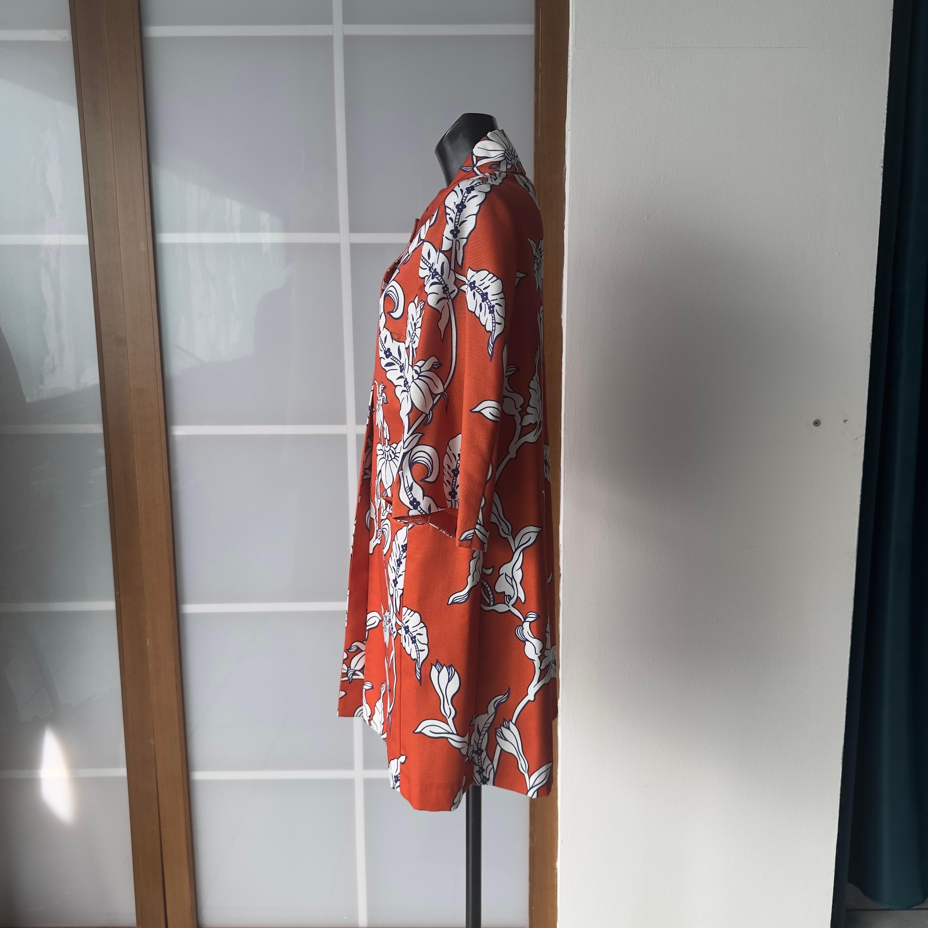 Spolverino Prada sui toni dell’arancio con stampa a fiori bianchi,  in ottime condizioni 
Taglio asimmetrico, scollo a giro e chiusura con bottoni automatici 
Due tasche frontali
Rimossa etichetta della composizione che presenta una rilevanza di