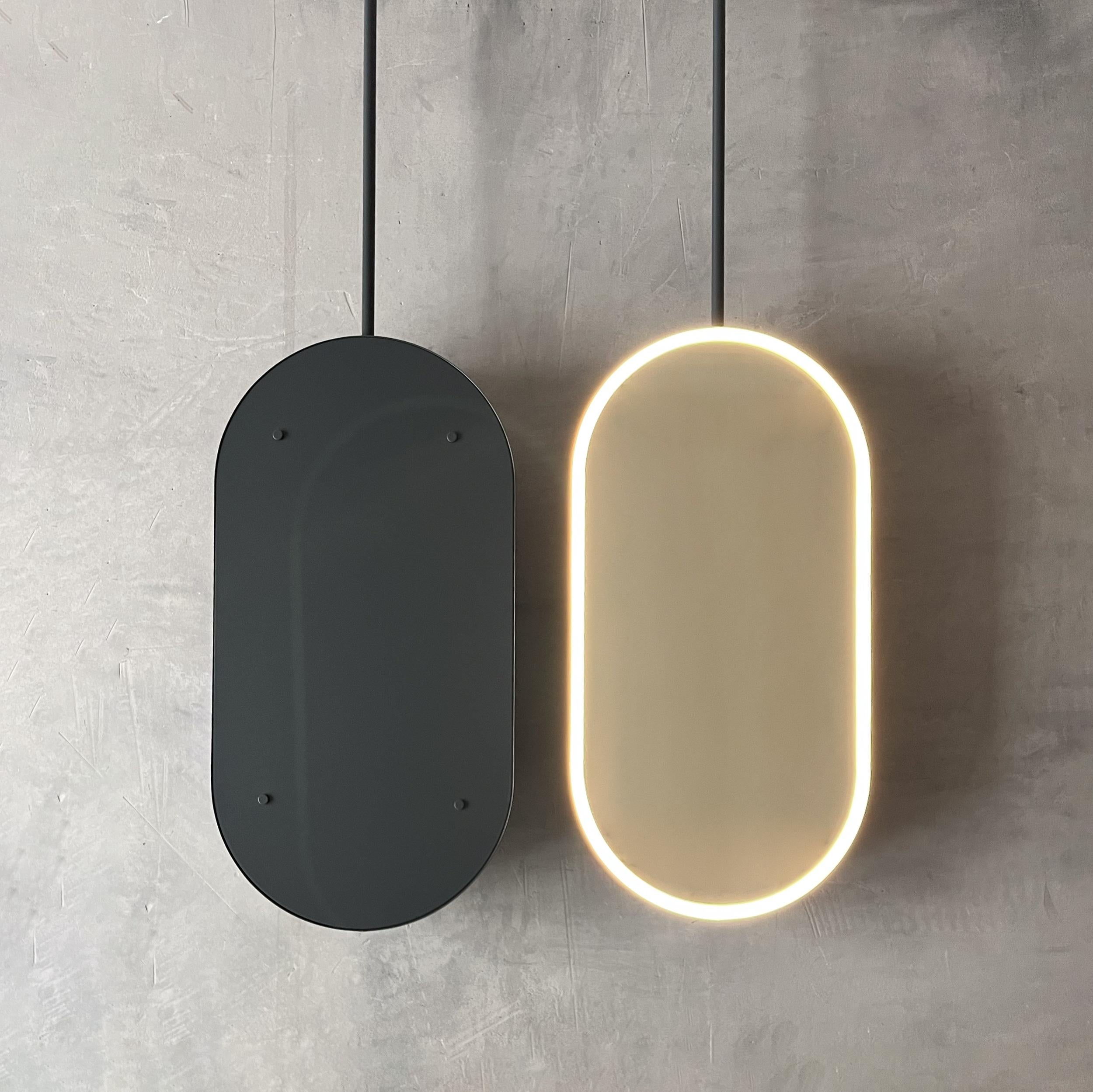 Contemporary front illuminated Capsula™ ceiling suspended pill shaped mirror with a minimalist powder coated anthracite grey frame, arm and backing. Designed and made in London, UK. This mirror is fully customisable to suit the specific requirements