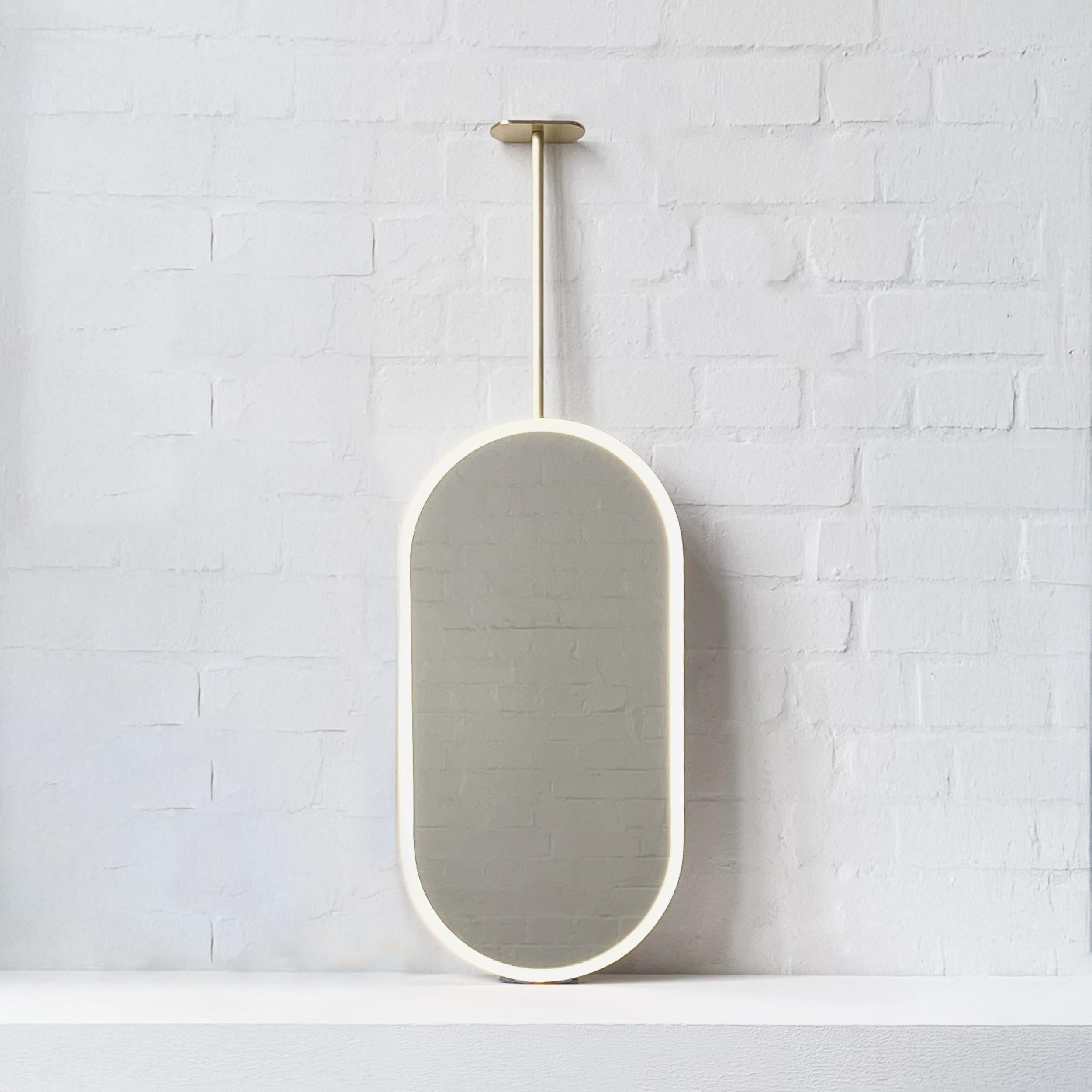 Moderno espejo en forma de píldora suspendido del techo Capsula™ con iluminación frontal y marco minimalista de latón cepillado. Diseñado y fabricado a mano en Londres, Reino Unido. Este espejo es totalmente personalizable para adaptarse a los