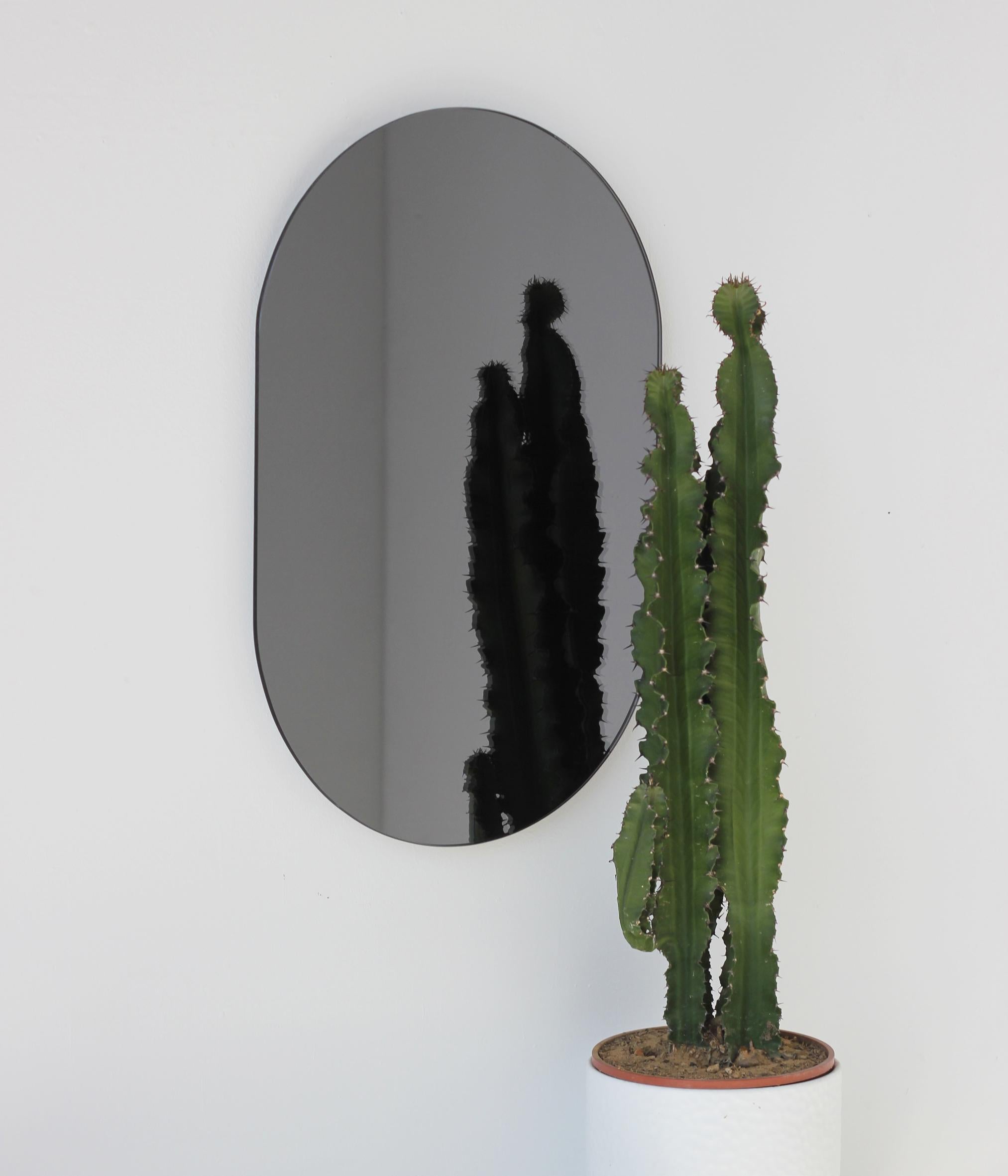 Minimalistischer, kapselförmiger, schwarz getönter, rahmenloser Spiegel. Hochwertiges Design, das dafür sorgt, dass der Spiegel perfekt parallel zur Wand steht. Entworfen und hergestellt in London, UK.

Ausgestattet mit professionellen Platten, die